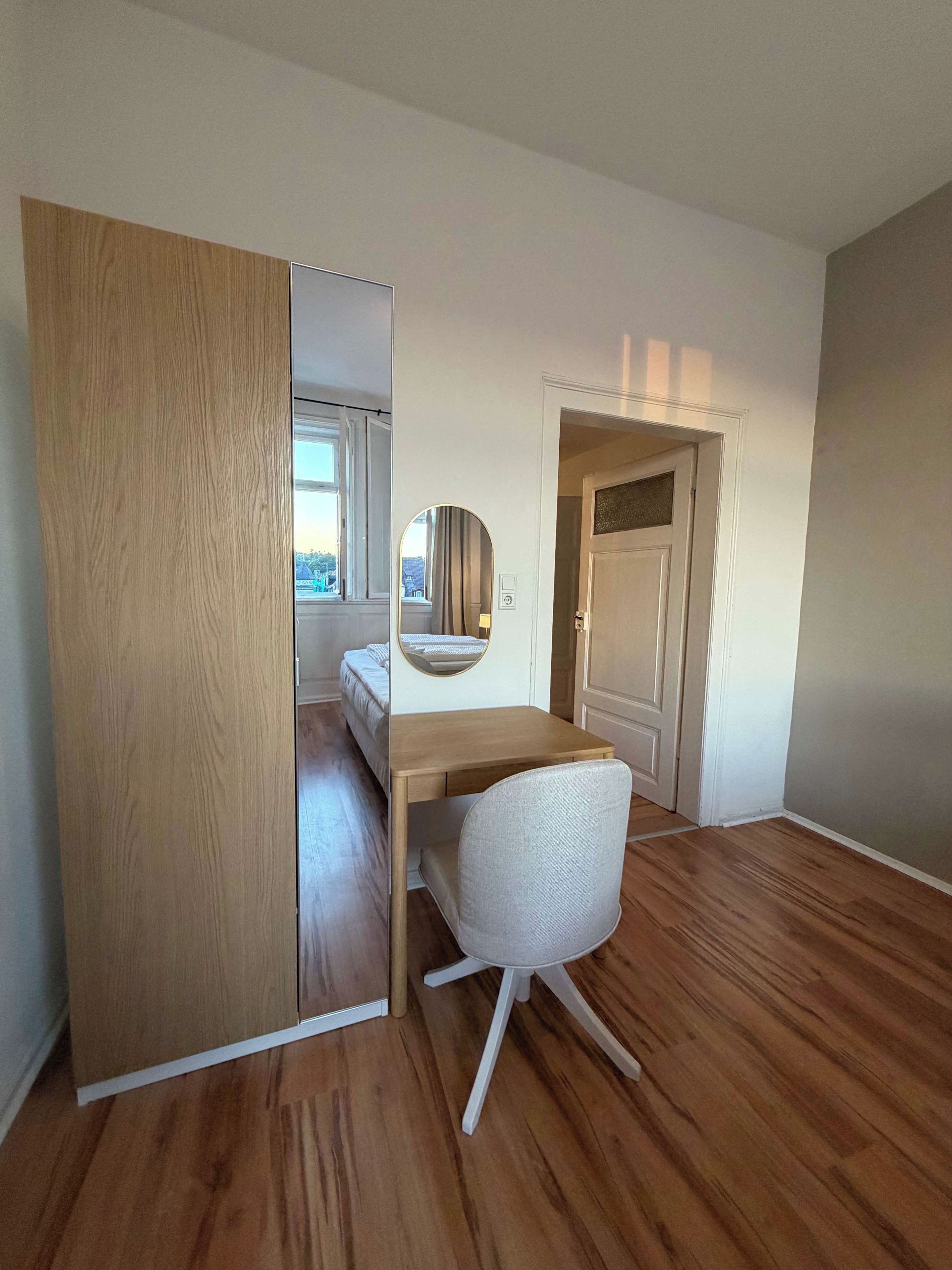 Une pièce avec un plancher en bois, un bureau avec une chaise, un miroir et une porte qui donne sur une autre pièce.