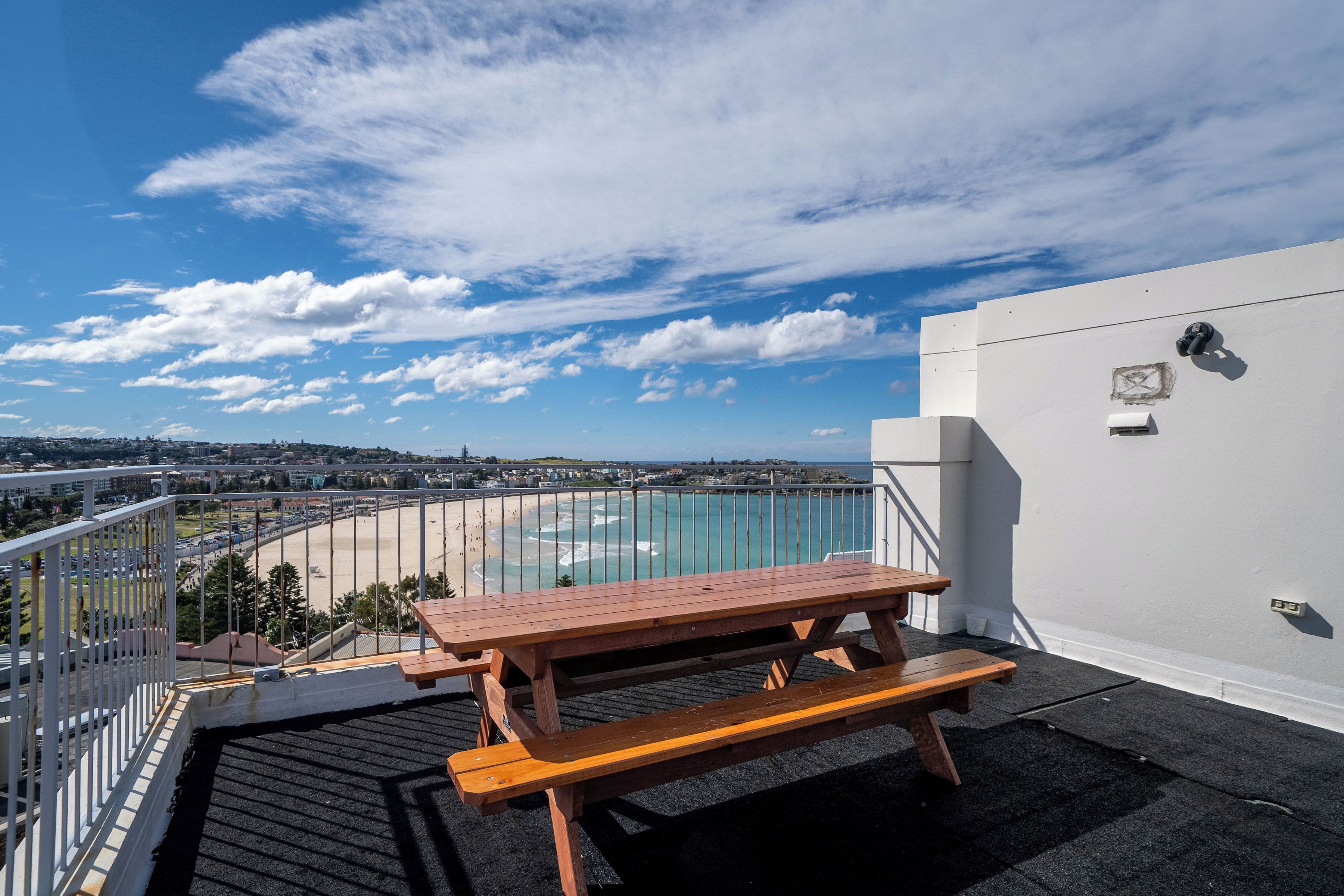 A wooden picnic table with benches on a rooftop terrace overlooking a beach and cityscape.