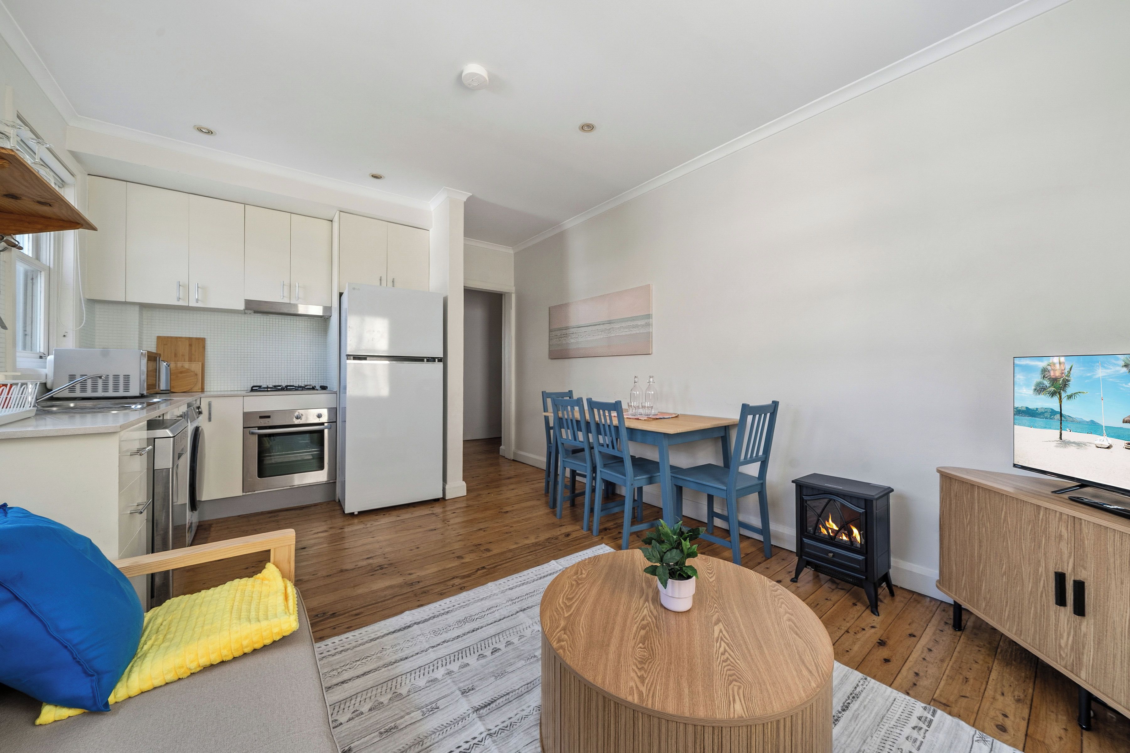 A modern kitchen with white appliances, a dining area with blue chairs, and a TV displaying a beach scene.