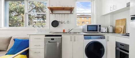 A compact kitchen with a dishwasher, microwave, and oven, a washing machine, and a window with a view of buildings and trees.