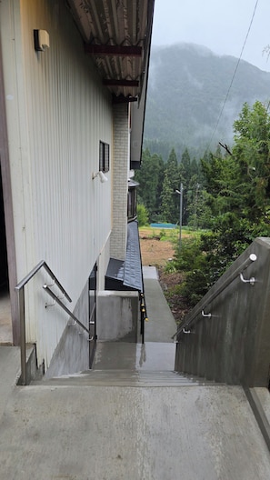 Una escalera de concreto que lleva a un edificio con barandilla de metal, un ventanal pequeño y vista a un paisaje verde con árboles y una colina al fondo.