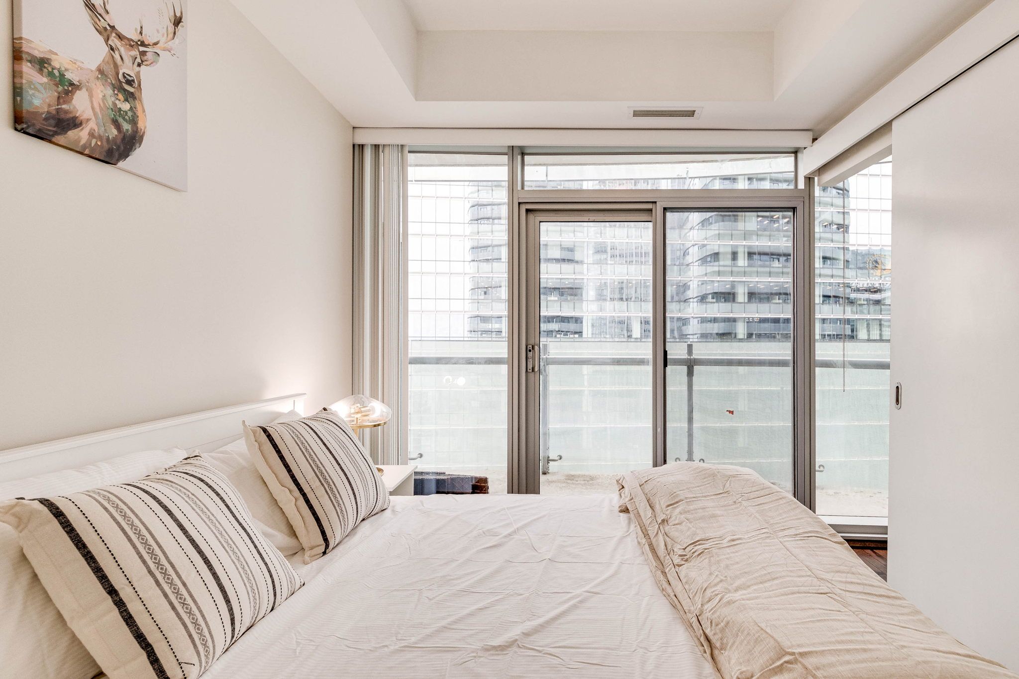 A modern bedroom with a large bed, a nightstand, a window with a view of buildings, and a ceiling light.