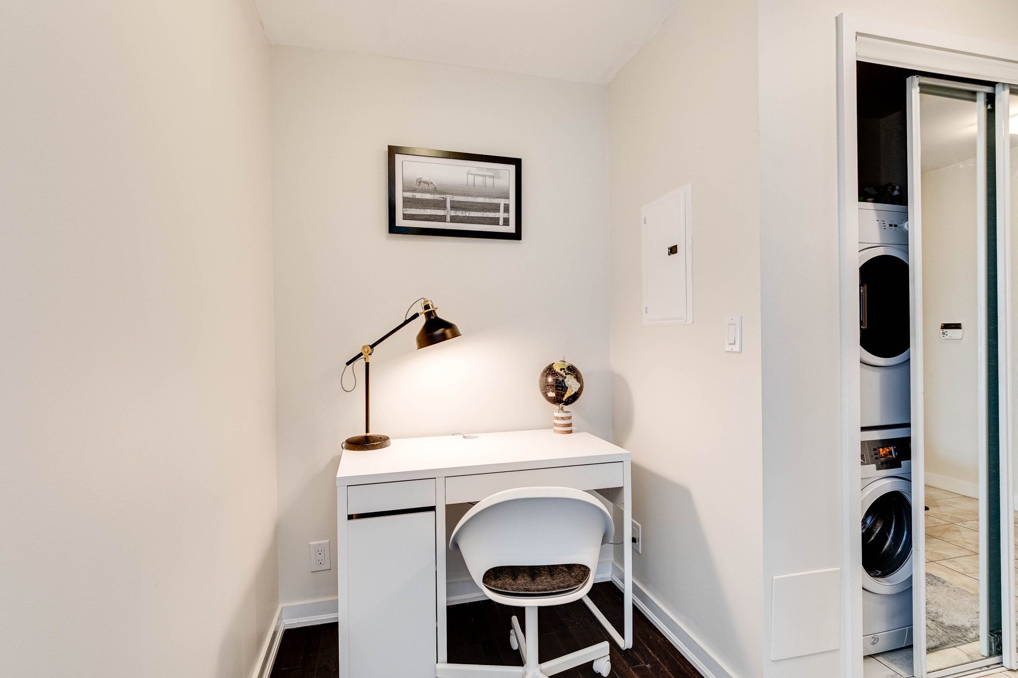 A compact room with a white desk, a chair, a desk lamp, a globe ornament, and a framed picture on the wall.