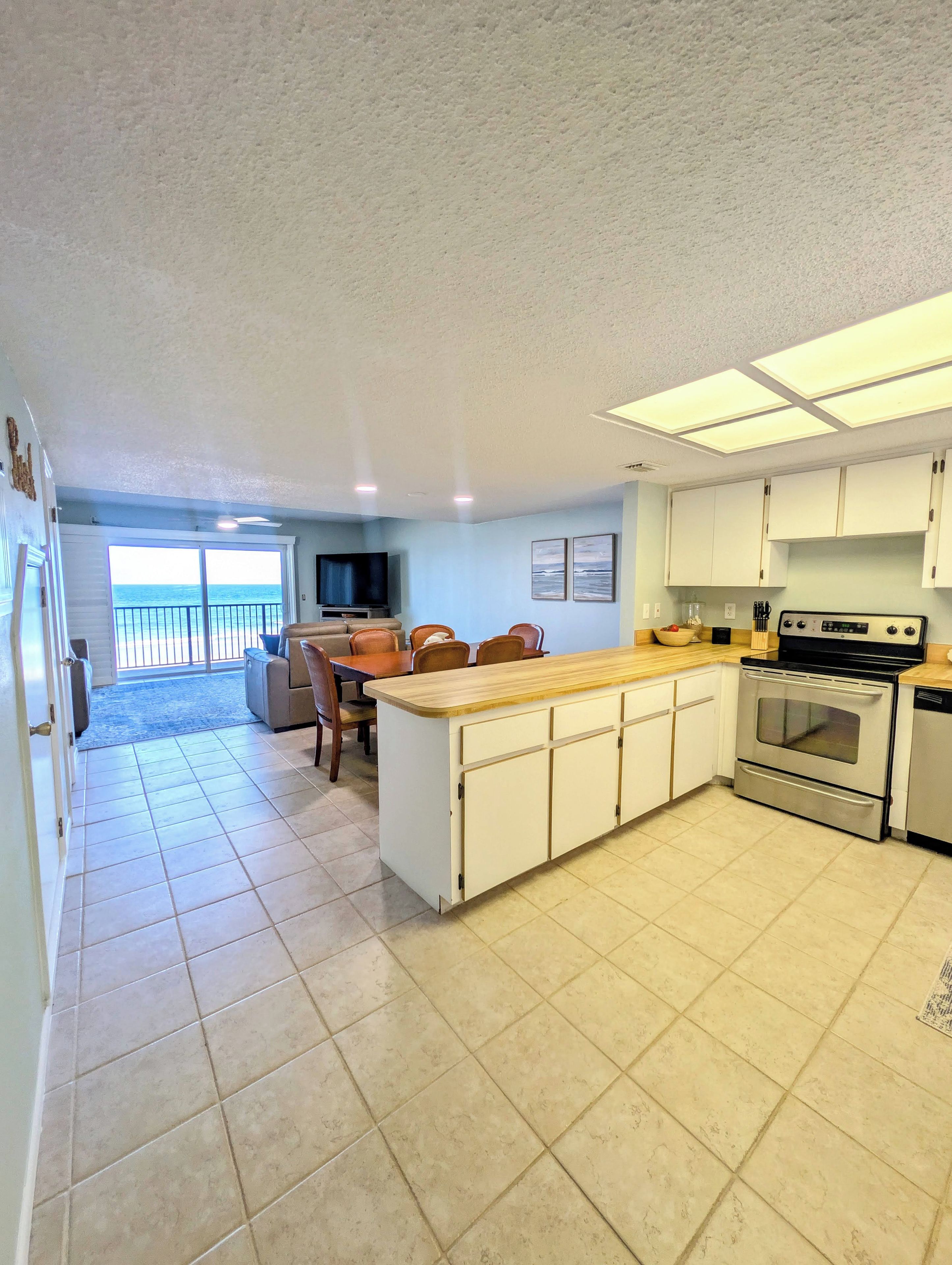 A kitchen with a central island, a dining area with chairs, and a view of the ocean through a sliding door.
