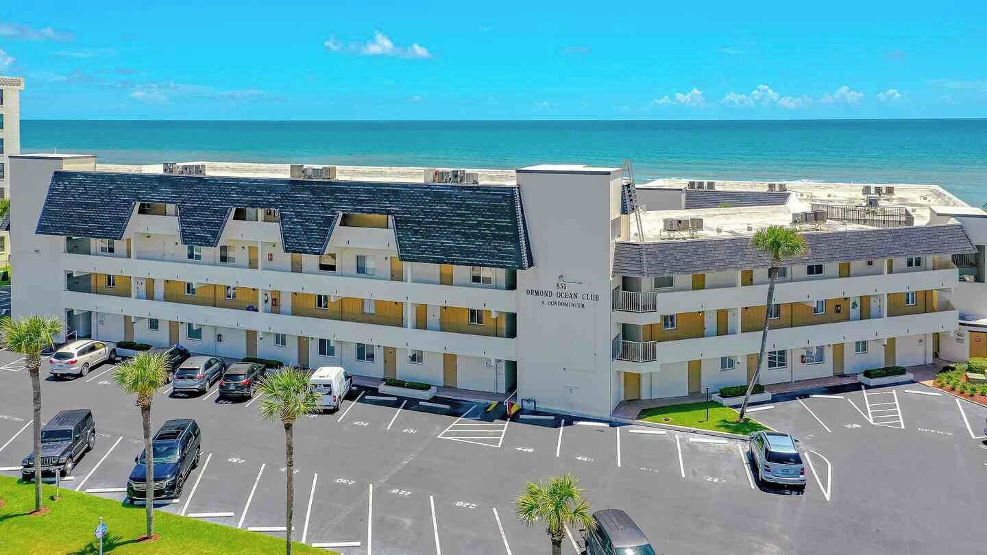Een meerhoog hotel met een parkeerplaats ervoor, uitzicht op het strand en een bord waarop staat dat het een resort is.