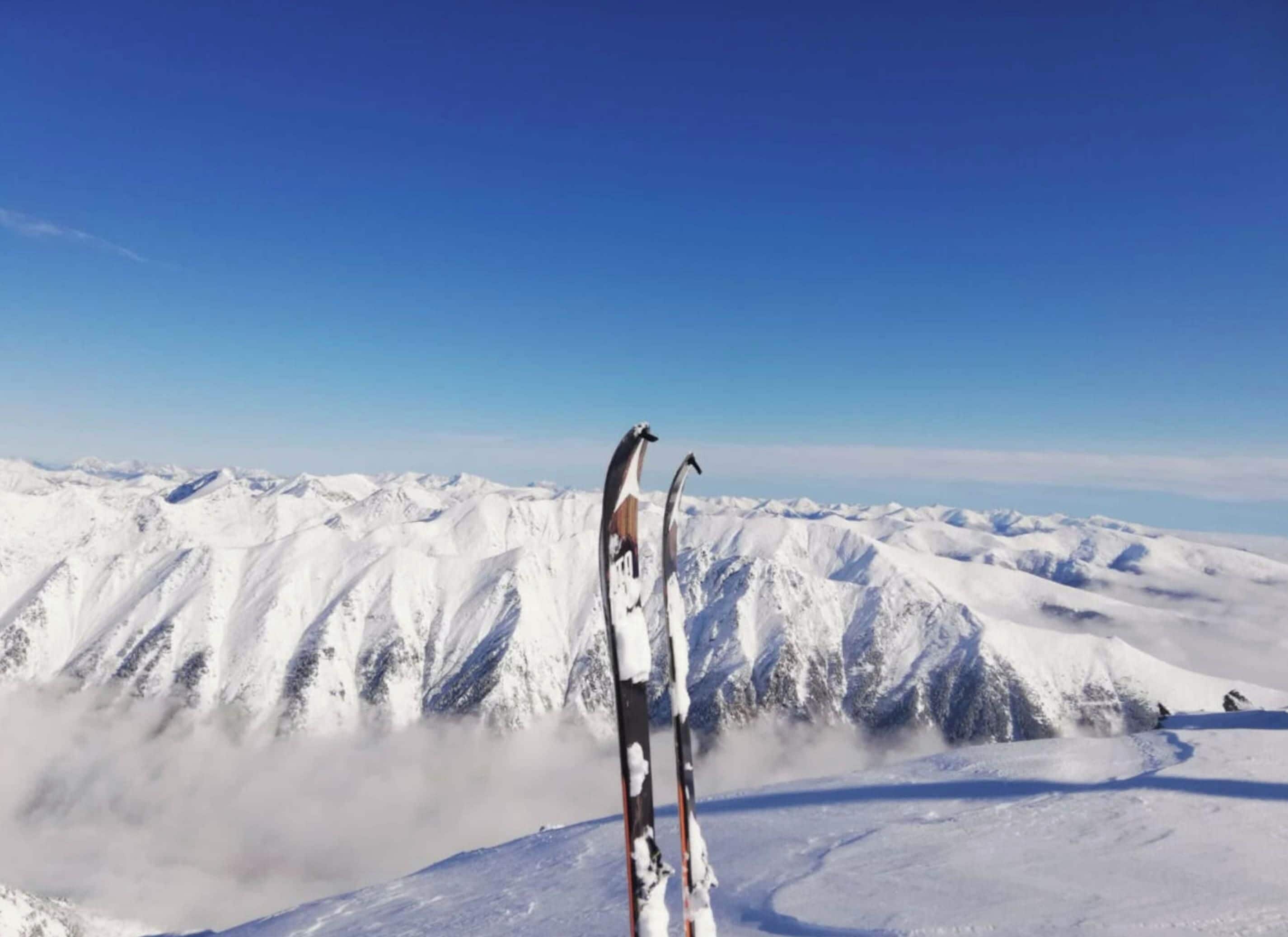 Ein Paar Ski steht aufstrebend auf einem verschneiten Berggipfel, über ihnen ein klarer Himmel.