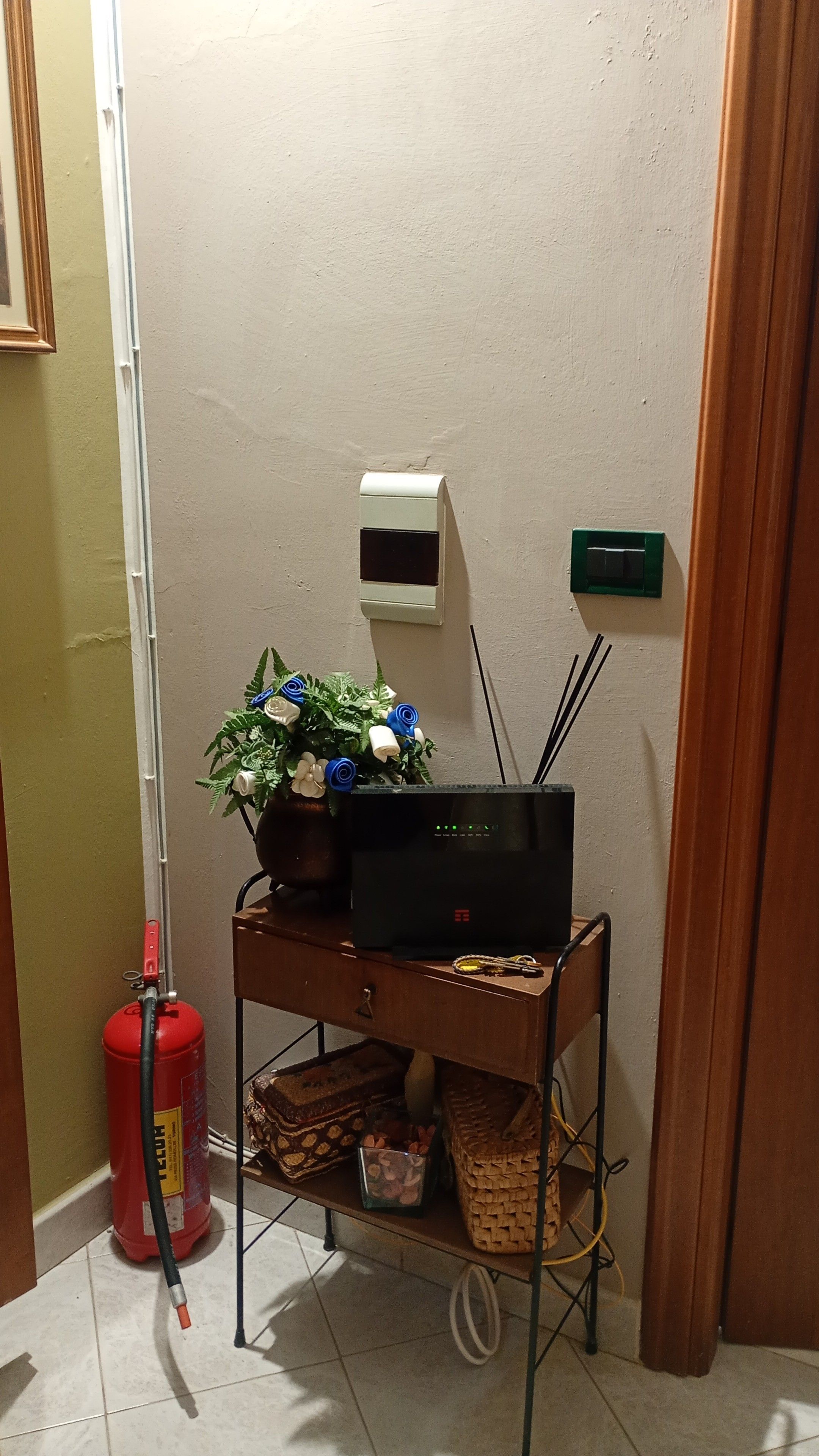 A room with a black flat-screen TV on a metal stand, a fire extinguisher, a wall-mounted air conditioning unit, and a small green sign.
