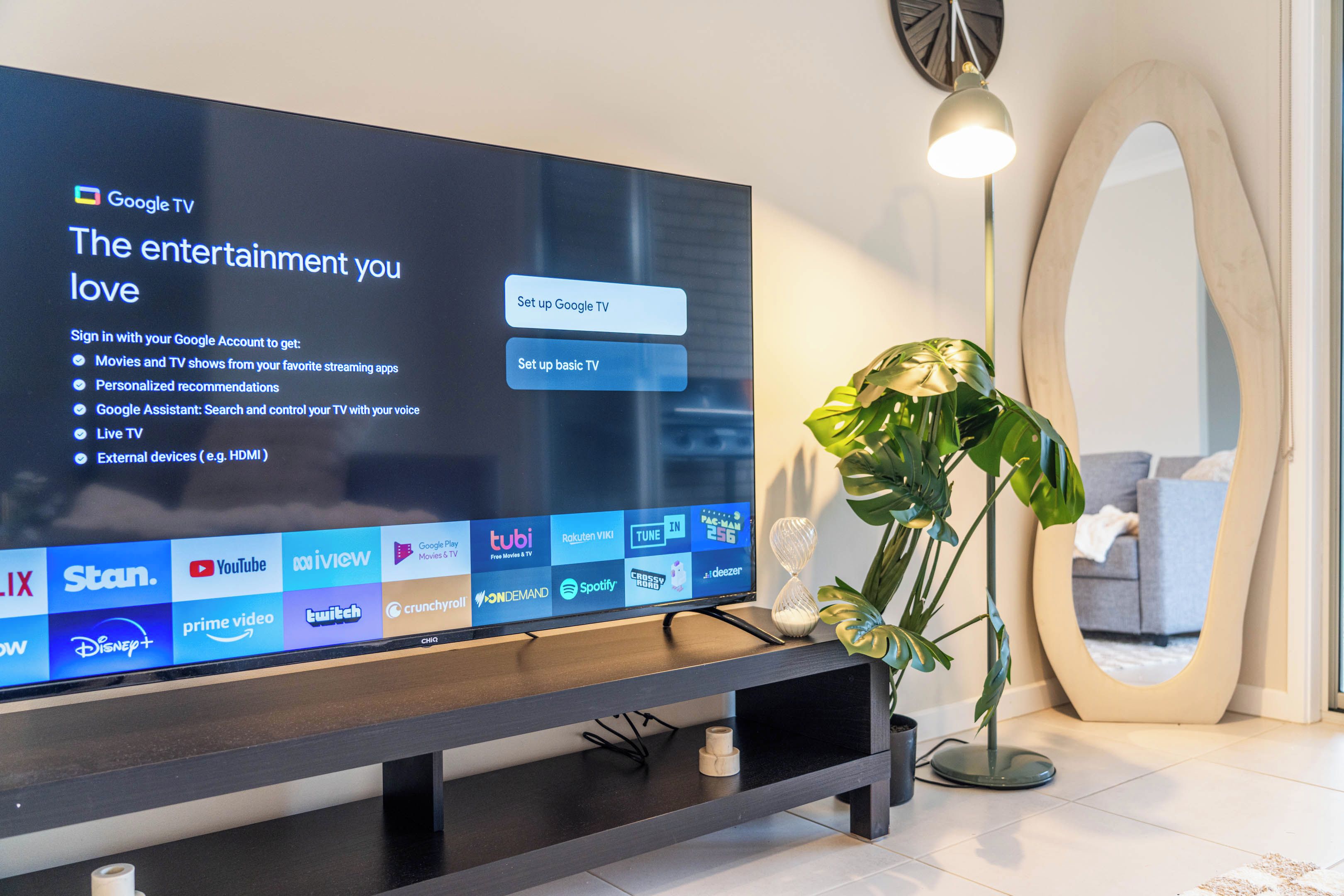 A flat-screen TV on a stand with a modern interior design, including a potted plant and a decorative mirror.