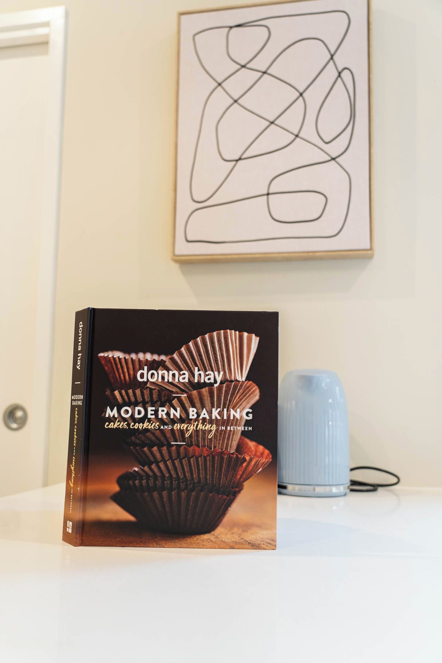 A book titled 'Modern Baking' by Donna Hay on a white surface with a blue vase nearby.