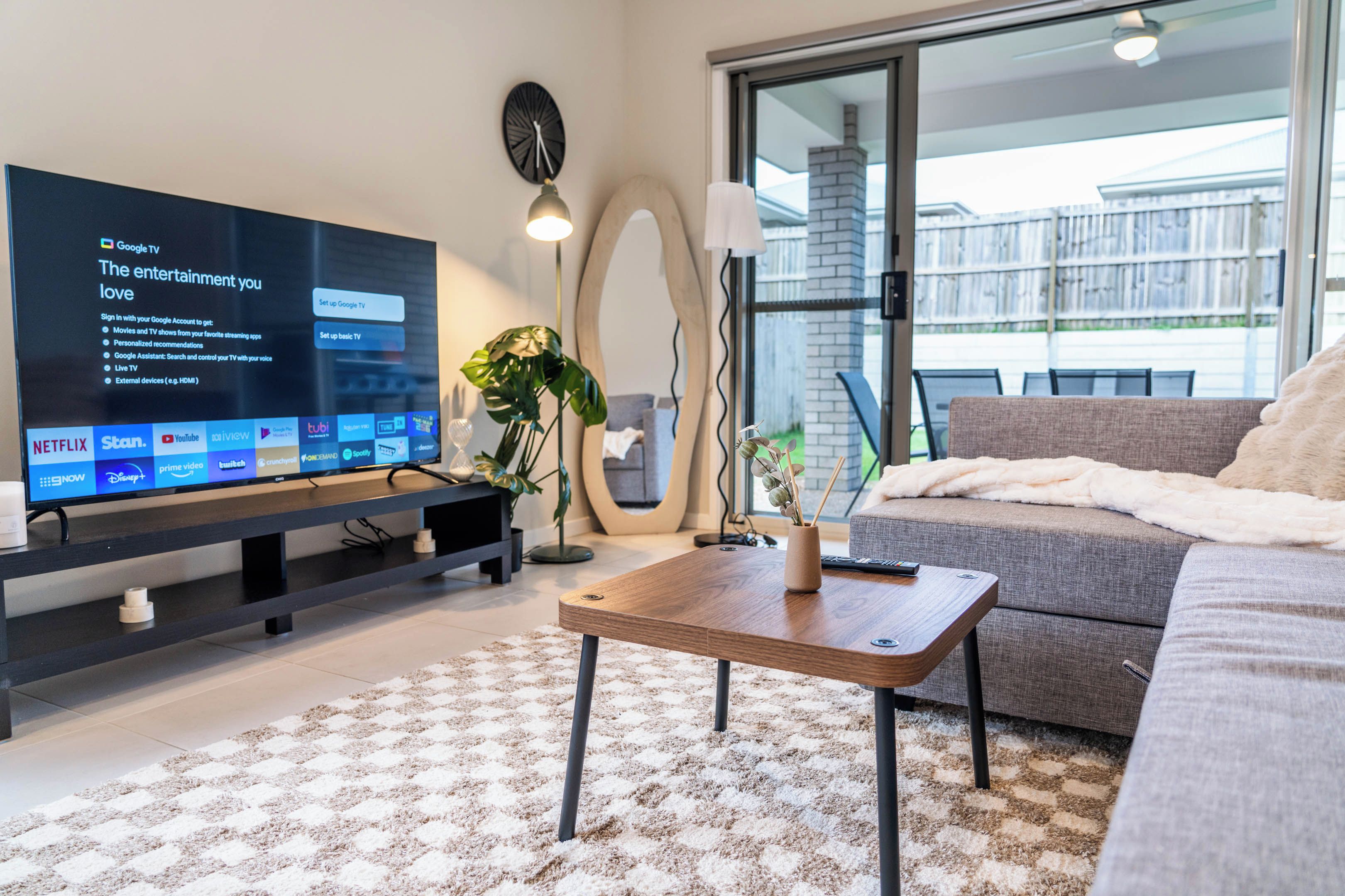 A modern living room with a flat-screen TV displaying streaming services, a sofa, a coffee table, and a large window with a view of a building.