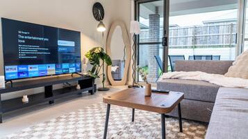 A modern living room with a flat-screen TV displaying streaming services, a sofa, a coffee table, and a large window with a view of a building.