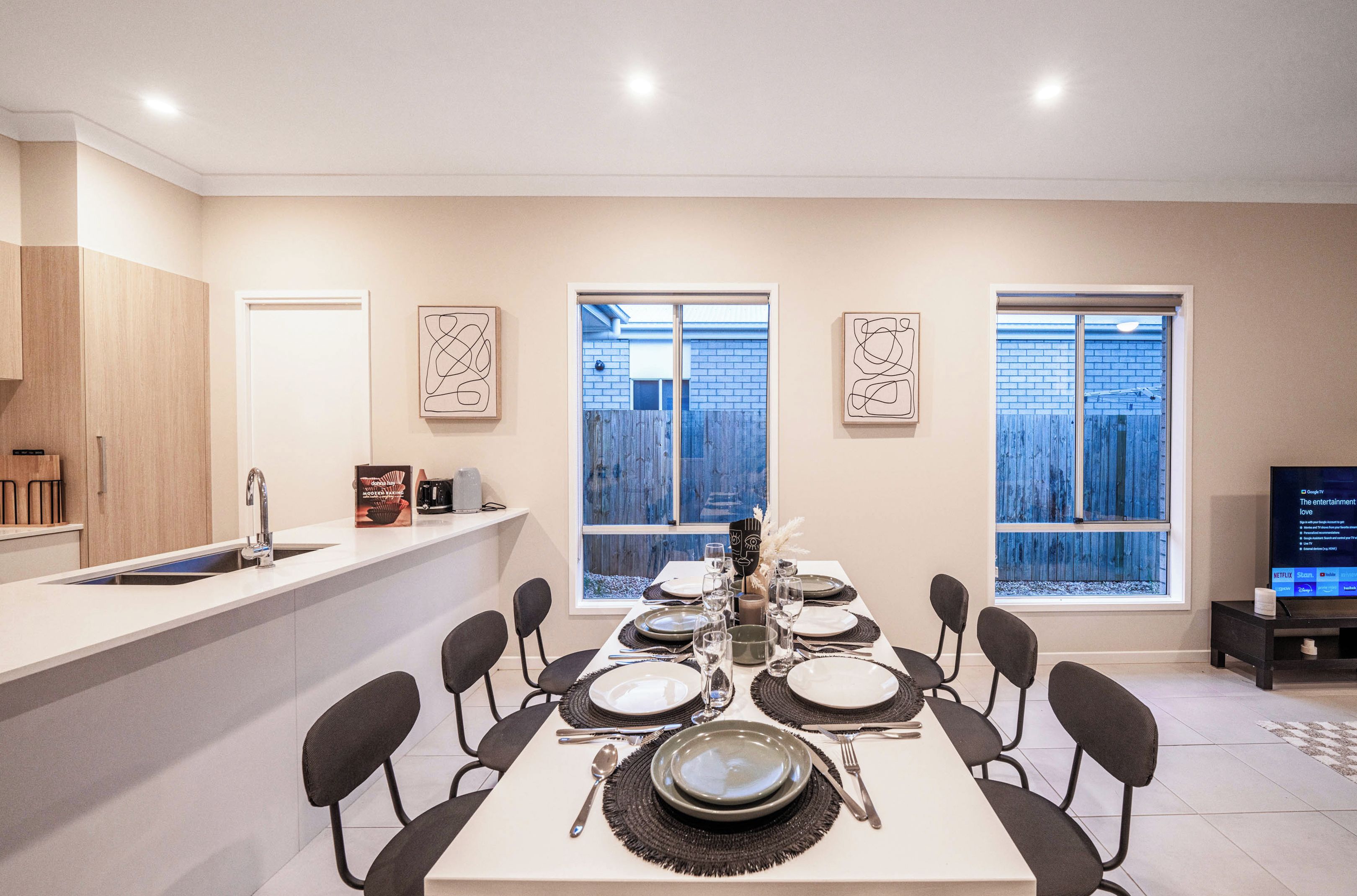 A dining area with a table set for six, a kitchen counter, and a television on a stand.