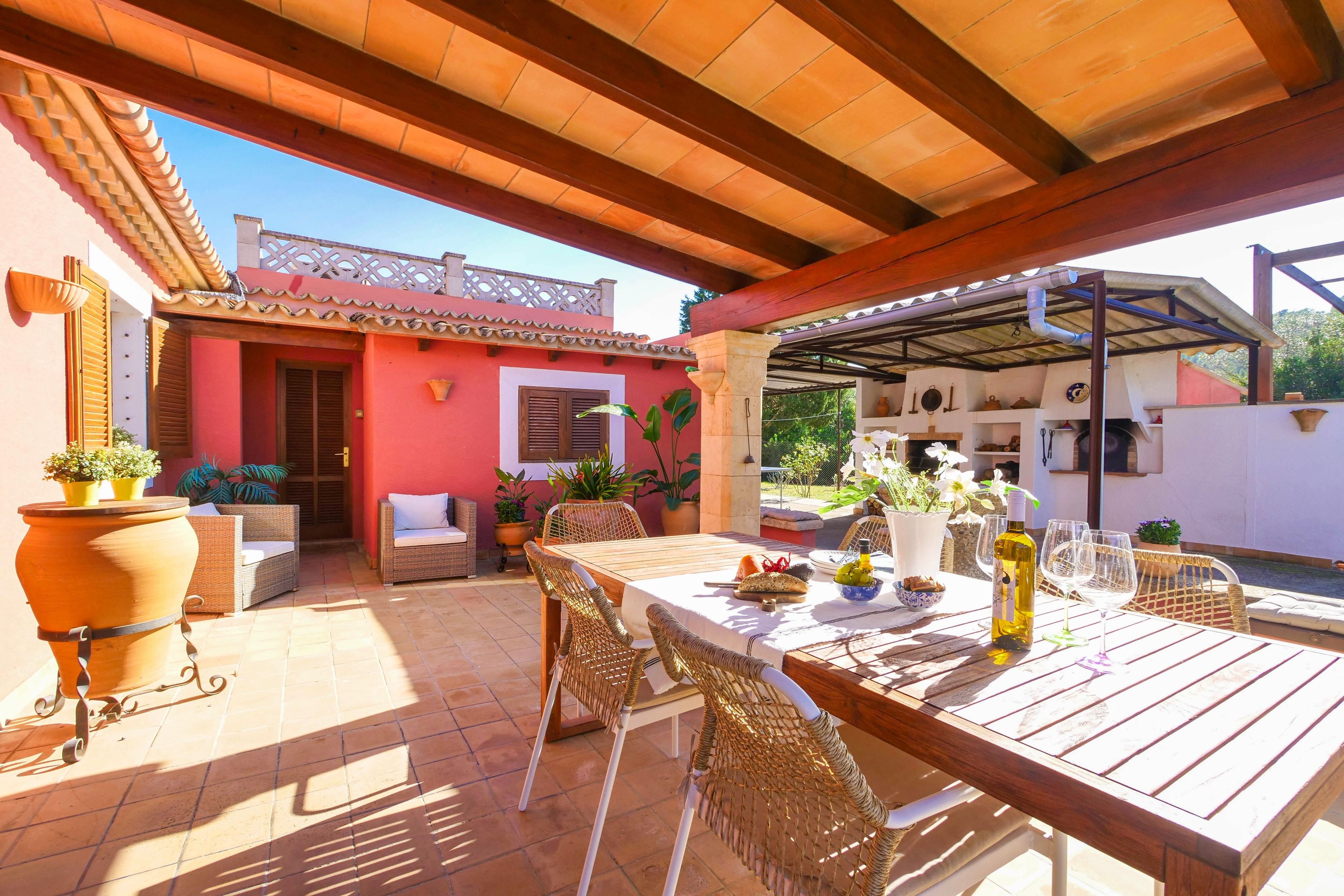 Un patio cubierto con una mesa de madera lista para comer, rodeado de sillas de mimbre, y un edificio rojo al fondo.