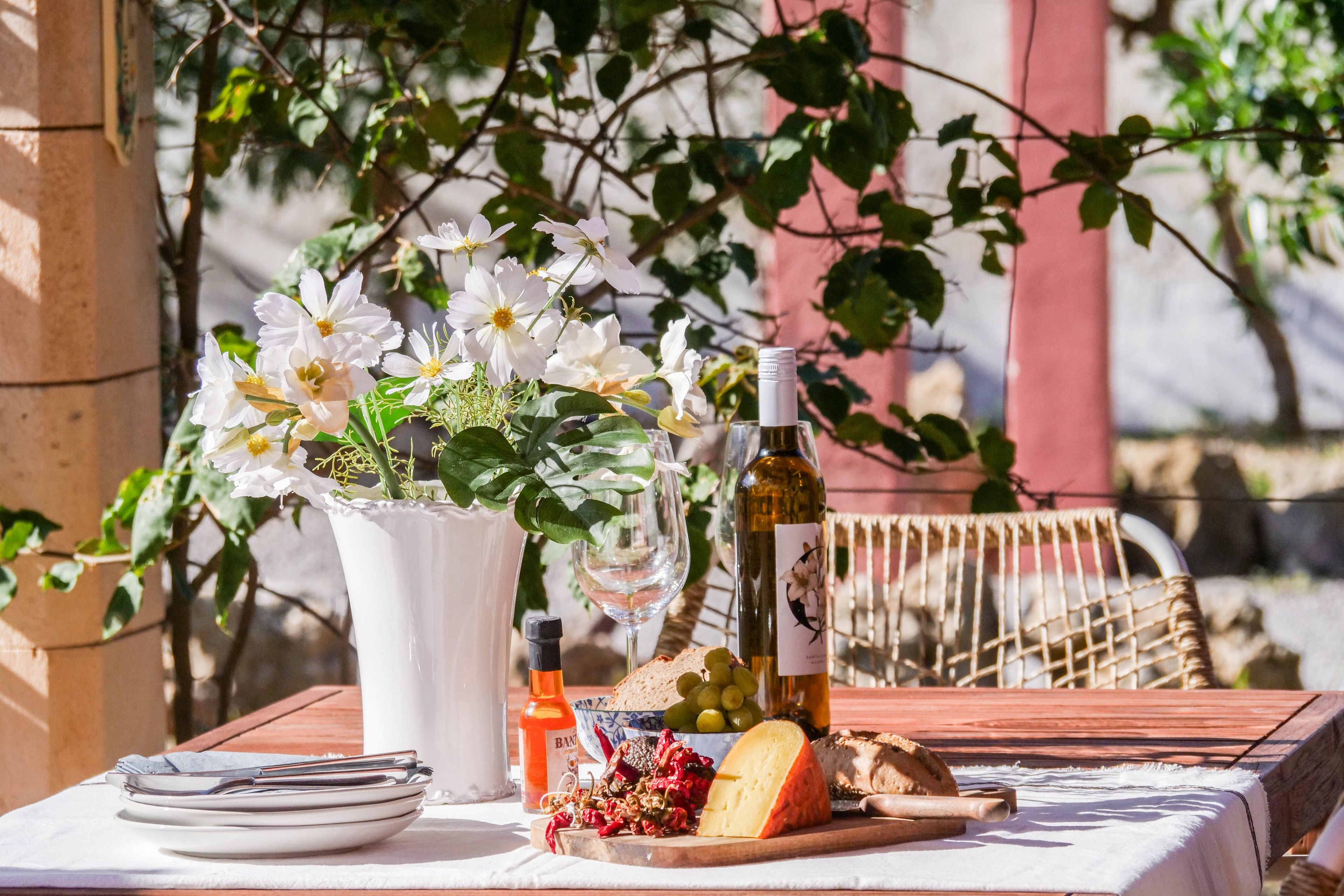 Una mesa servida con platos, una botella, una copa, una botella de vino, un bol de uvas, un trozo de queso y un jarrón con flores.