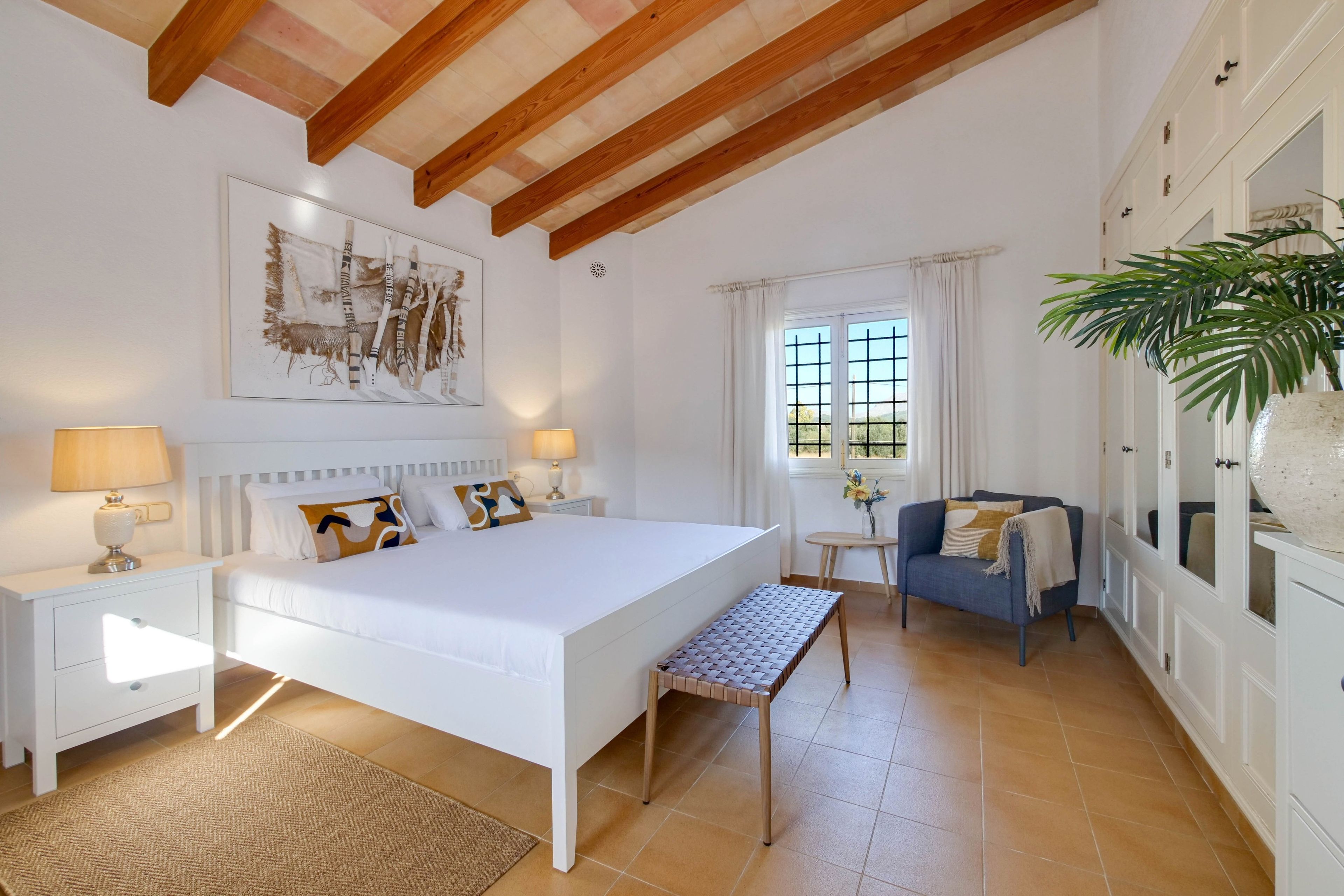 Un dormitorio con una cama grande, mesitas de noche, un banco, un sofá, una ventana con cortinas y una planta.