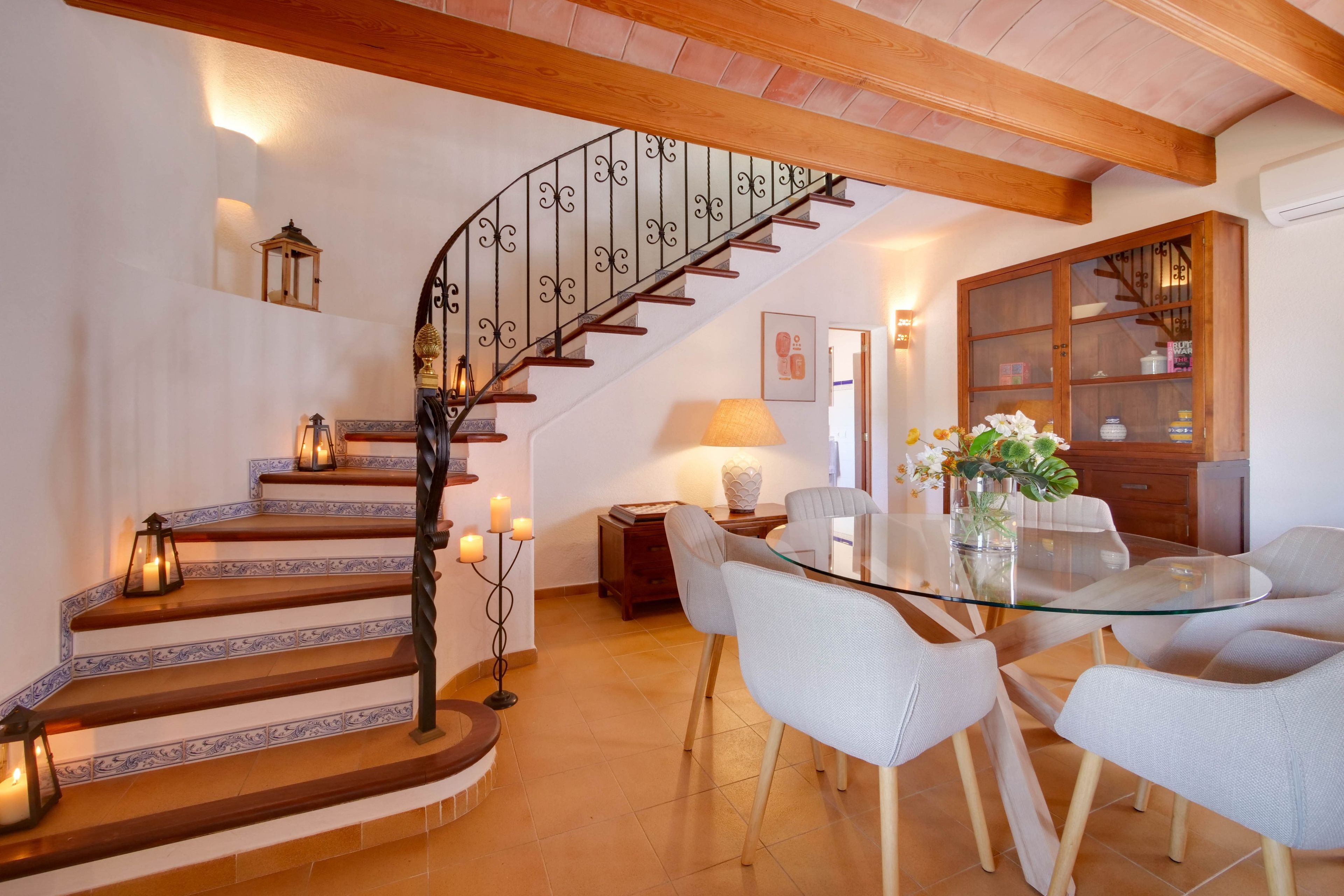 Un comedor con mesa de vidrio rodeada de sillas, una escalera con barandilla de hierro forjado y un mueble de madera con puertas de vidrio.
