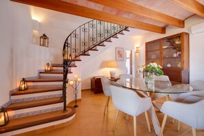 A dining area with a glass table surrounded by chairs, a staircase with a wrought iron railing, and a wooden cabinet with glass doors. - Finca Reig – Vacation Home with Private Pool near Pollensa Beach (Illes Balears)