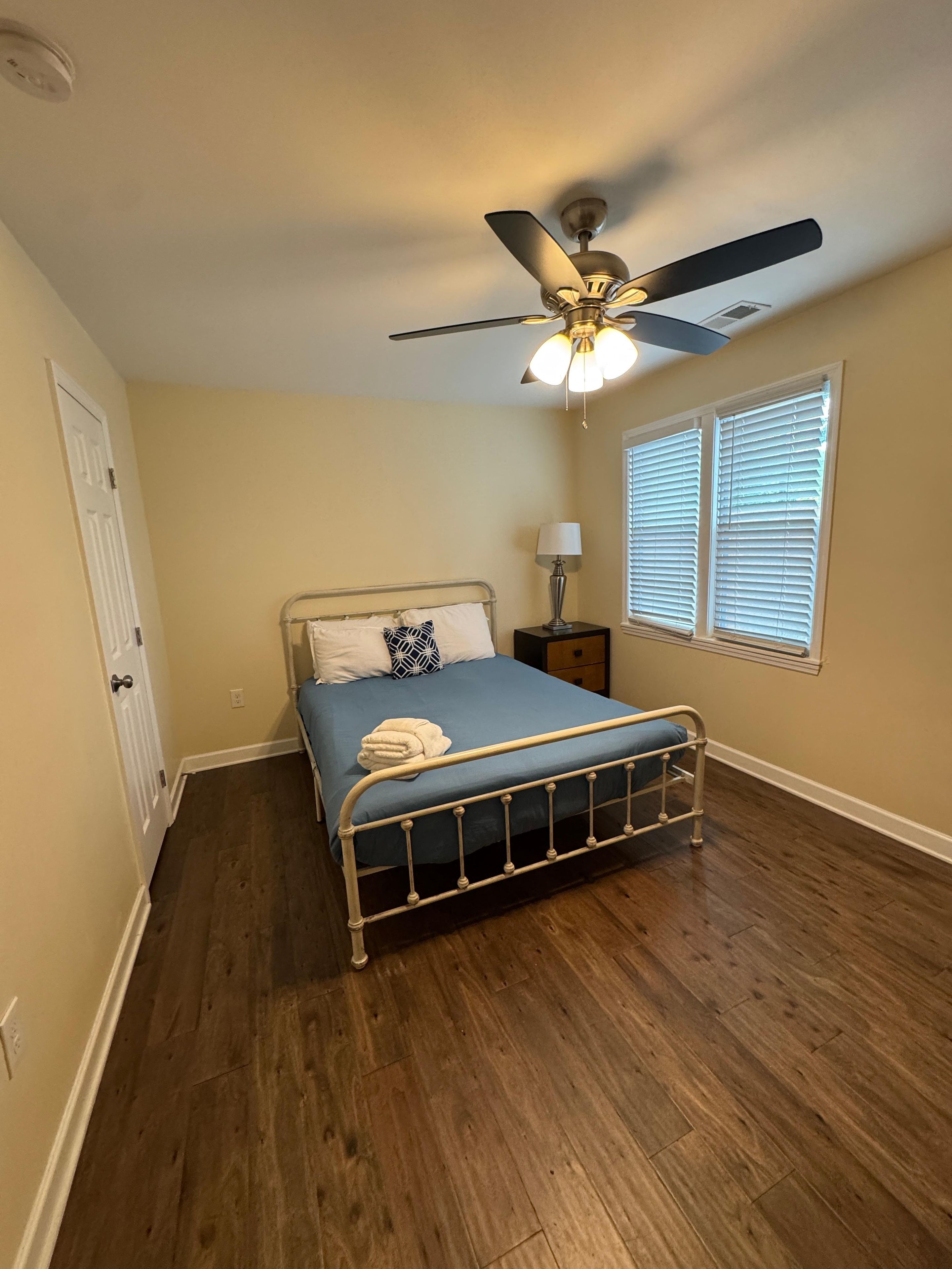 A bedroom with a bed, a ceiling fan, a window with blinds, and a door.