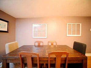 Un coin repas avec une table en bois et quatre chaises, deux fenêtres à cadres blancs et un tableau encadré au mur.