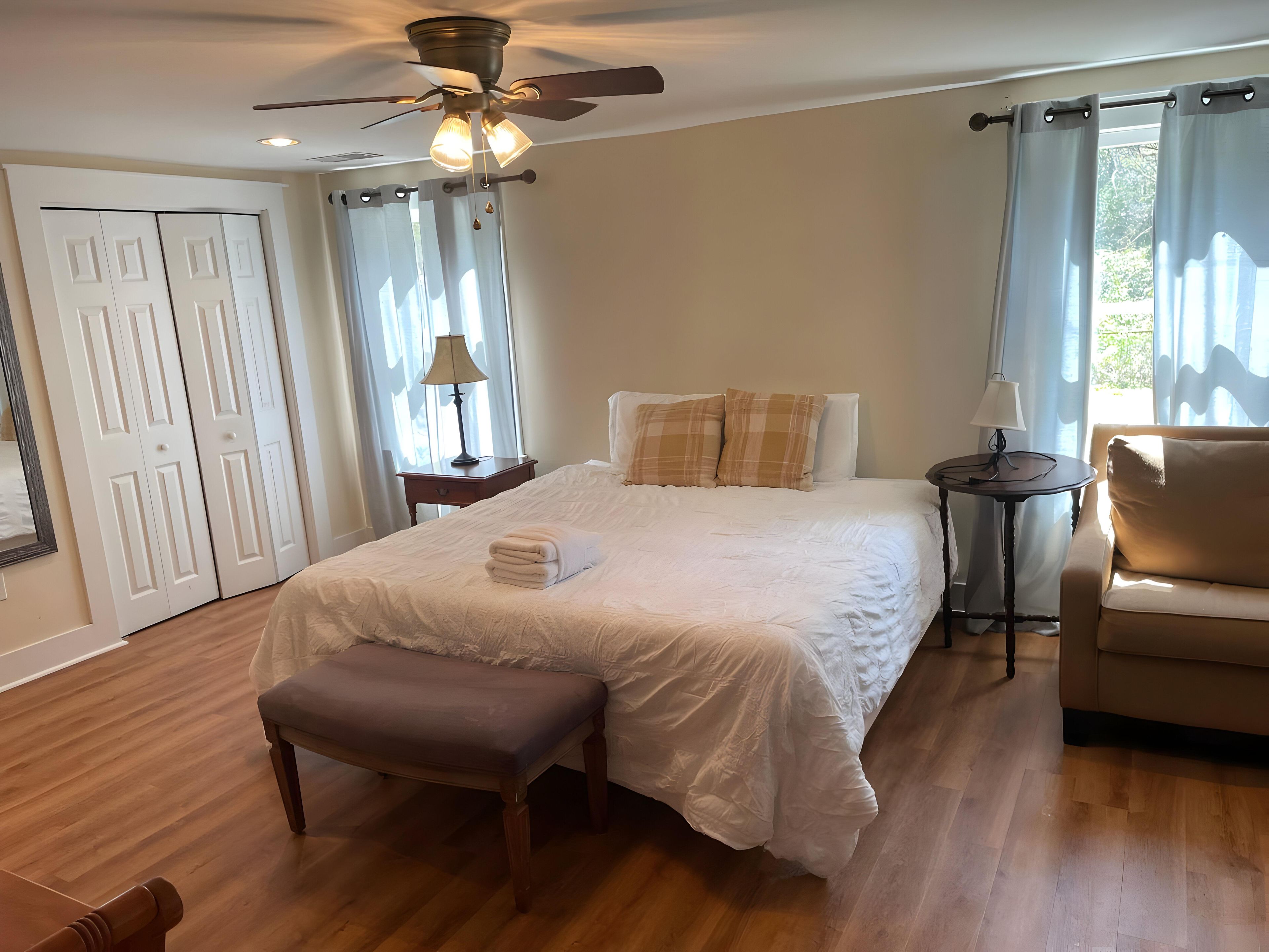 A bedroom with a bed, a bench, a nightstand, a chair, a ceiling fan, and a window with curtains.