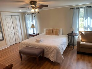 A bedroom with a bed, a bench, a nightstand, a chair, a ceiling fan, and a window with curtains. - Janelle’s Cottage: Walk to Brewery+Relax on Patio! (North Charleston)