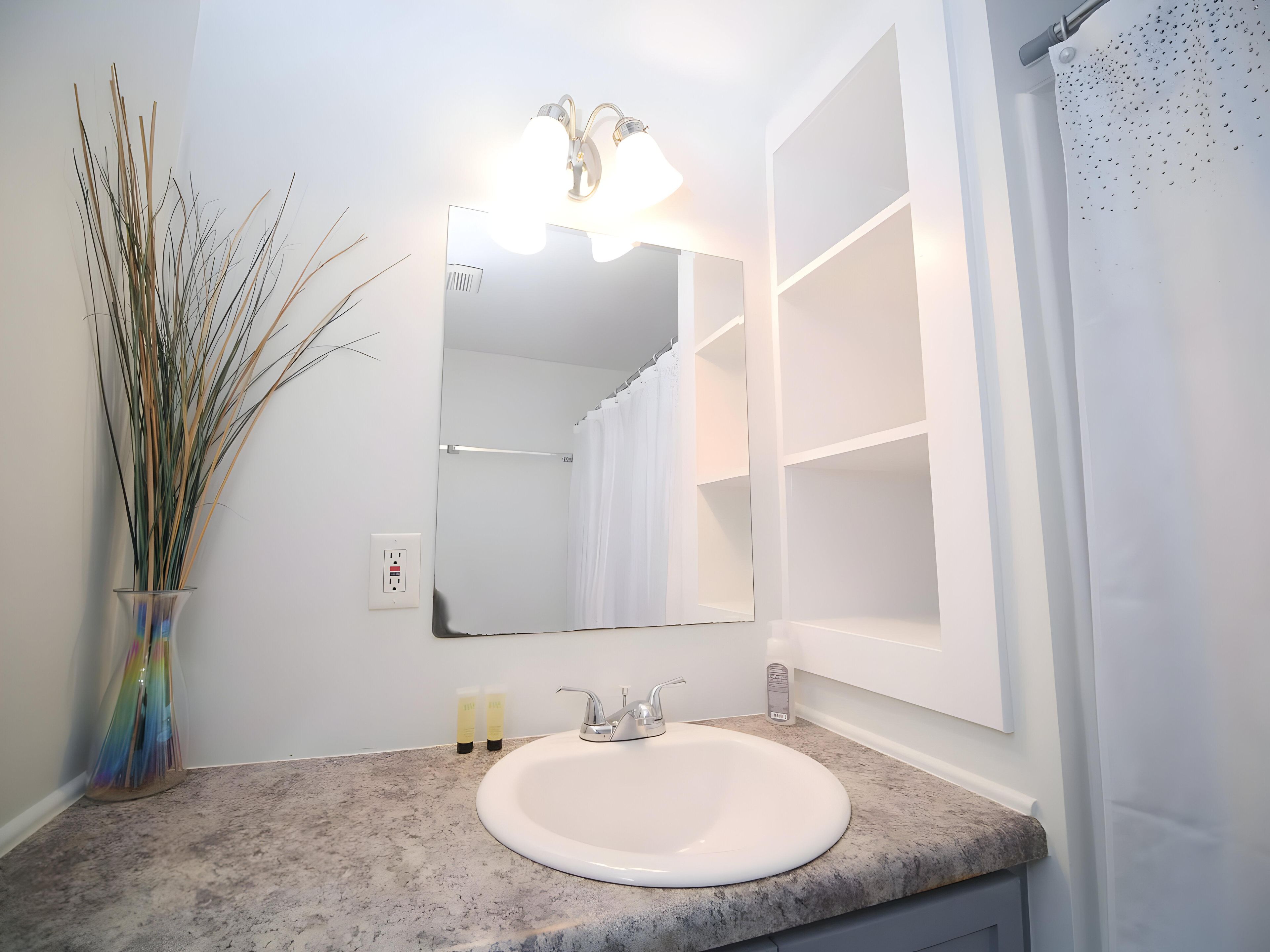 A bathroom with a white sink, a mirror, a shower curtain, and a vase with reeds.