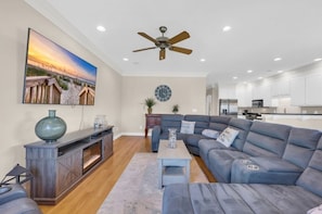 A modern living room with a sectional sofa, a flat-screen TV, a ceiling fan, and a kitchen in the background. - Steps to Beach + Private Pool | Sleeps 12 | 5BR (Murrells Inlet)