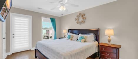 A bedroom with a bed, a nightstand, a ceiling fan, a window with curtains, and a view of buildings outside.