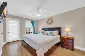 A bedroom with a bed, a nightstand, a ceiling fan, a window with curtains, and a view of buildings outside. - Steps to Beach + Private Pool | Sleeps 12 | 5BR (Murrells Inlet)