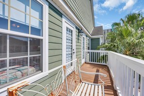 A balcony with a white railing, a metal chair, and a view of a building with large windows and a green exterior. - Steps to Beach + Private Pool | Sleeps 12 | 5BR (Murrells Inlet)