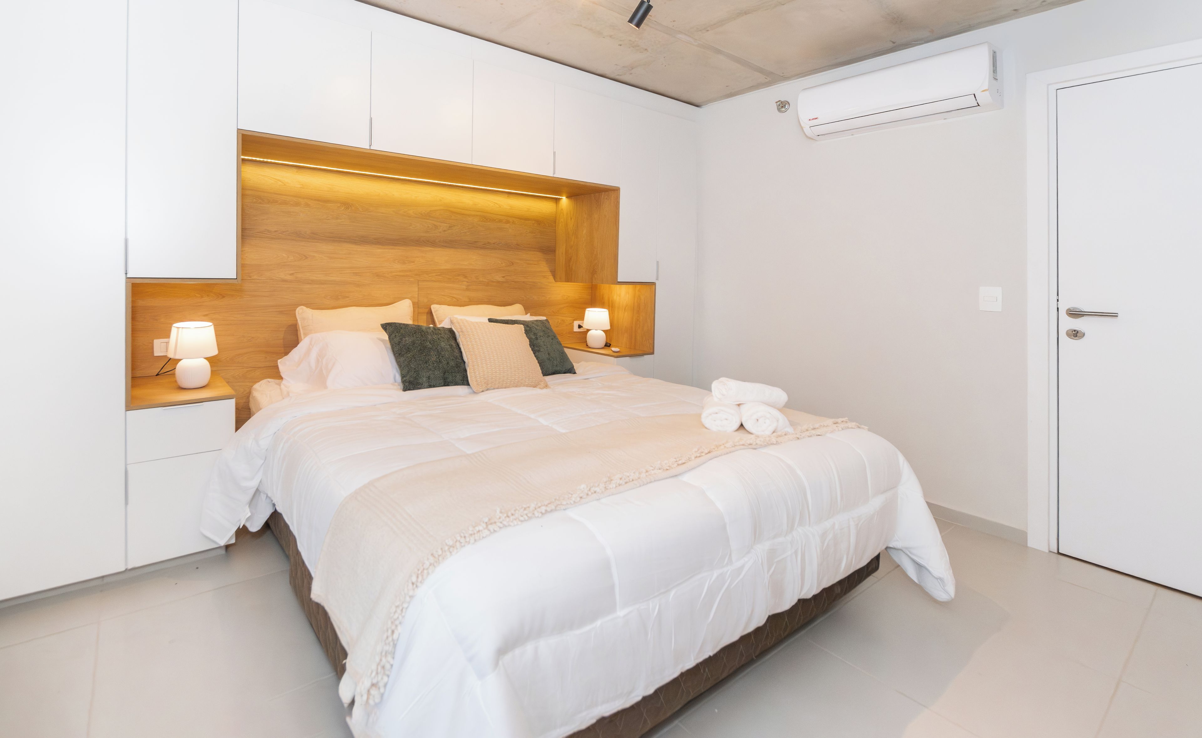 A hotel room with a bed, bedside lamps, a wooden headboard, a wall-mounted air conditioner, and a door.