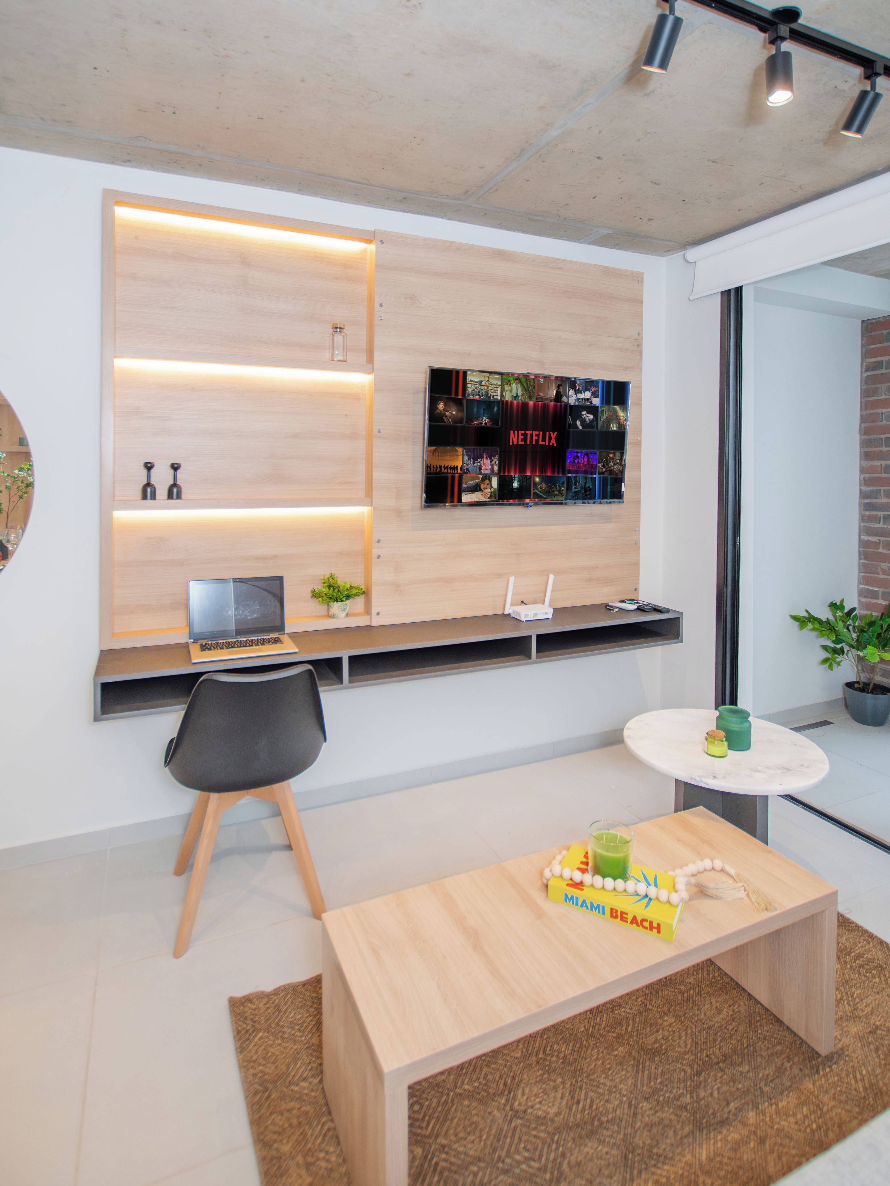 A modern room with a flat-screen TV mounted on a wooden wall, a desk with a laptop, a chair, and a small round table with a drink and a book.