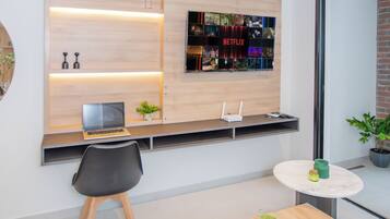 A modern room with a flat-screen TV mounted on a wooden wall, a desk with a laptop, a chair, and a small round table with a drink and a book.
