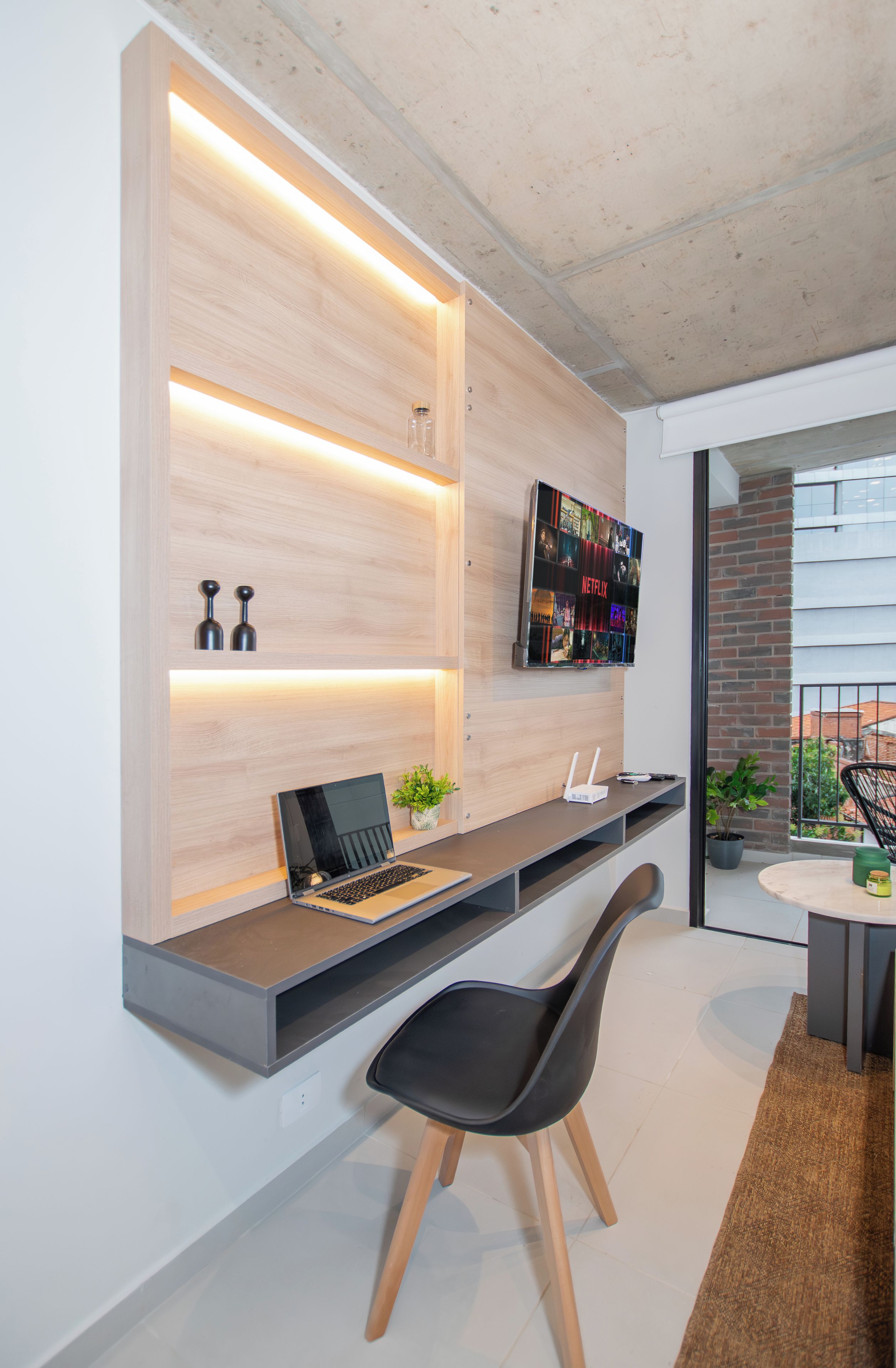 A modern home office with a built-in desk, a laptop, a plant, and a wall-mounted TV displaying the Netflix logo.