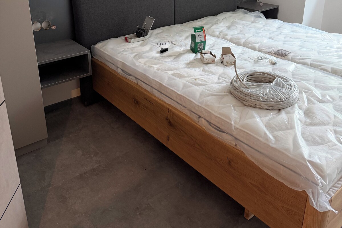 Una camera da letto con una struttura letto in legno, una testiera grigia, un comodino e una piccola mensola.