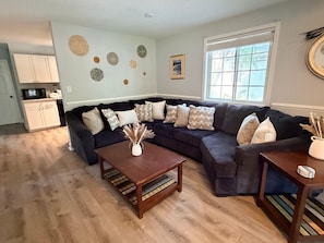 Ein Wohnzimmer mit einem dunkelgrauen Ecksofa, einem Holztisch und einem Esstisch mit Stühlen. Es gibt eine Küchenzeile mit weißen Schränken und einer Mikrowelle. Der Raum verfügt über Holzböden und ein großes Fenster mit Jalousien.