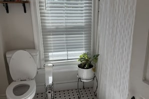 Ein Badezimmer mit Toilette, einem Fenster mit Jalousien und einer Topfpflanze auf einem kleinen Beistelltisch.