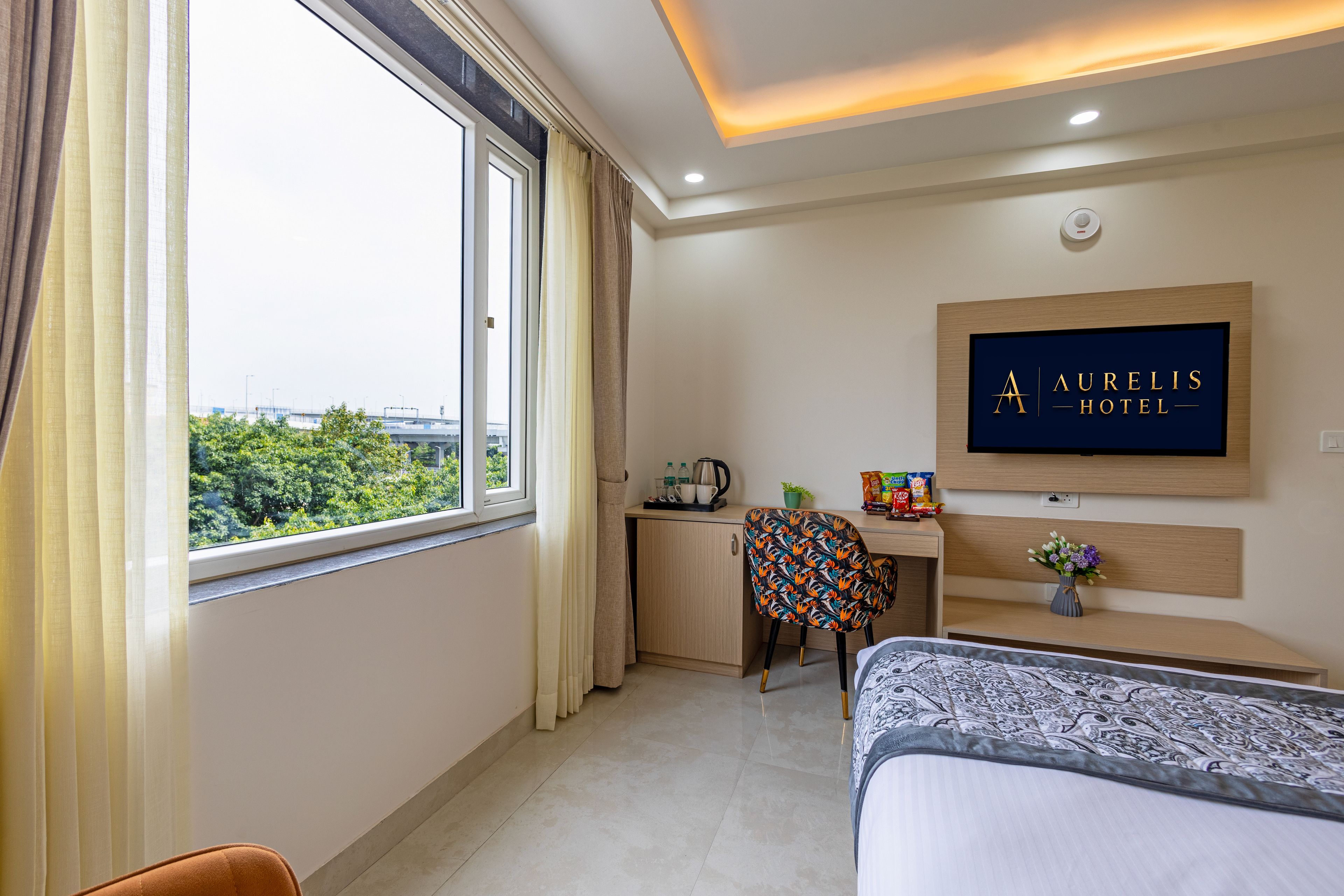 A hotel room with a bed, a desk, a chair, a small kitchenette, and a window with curtains.