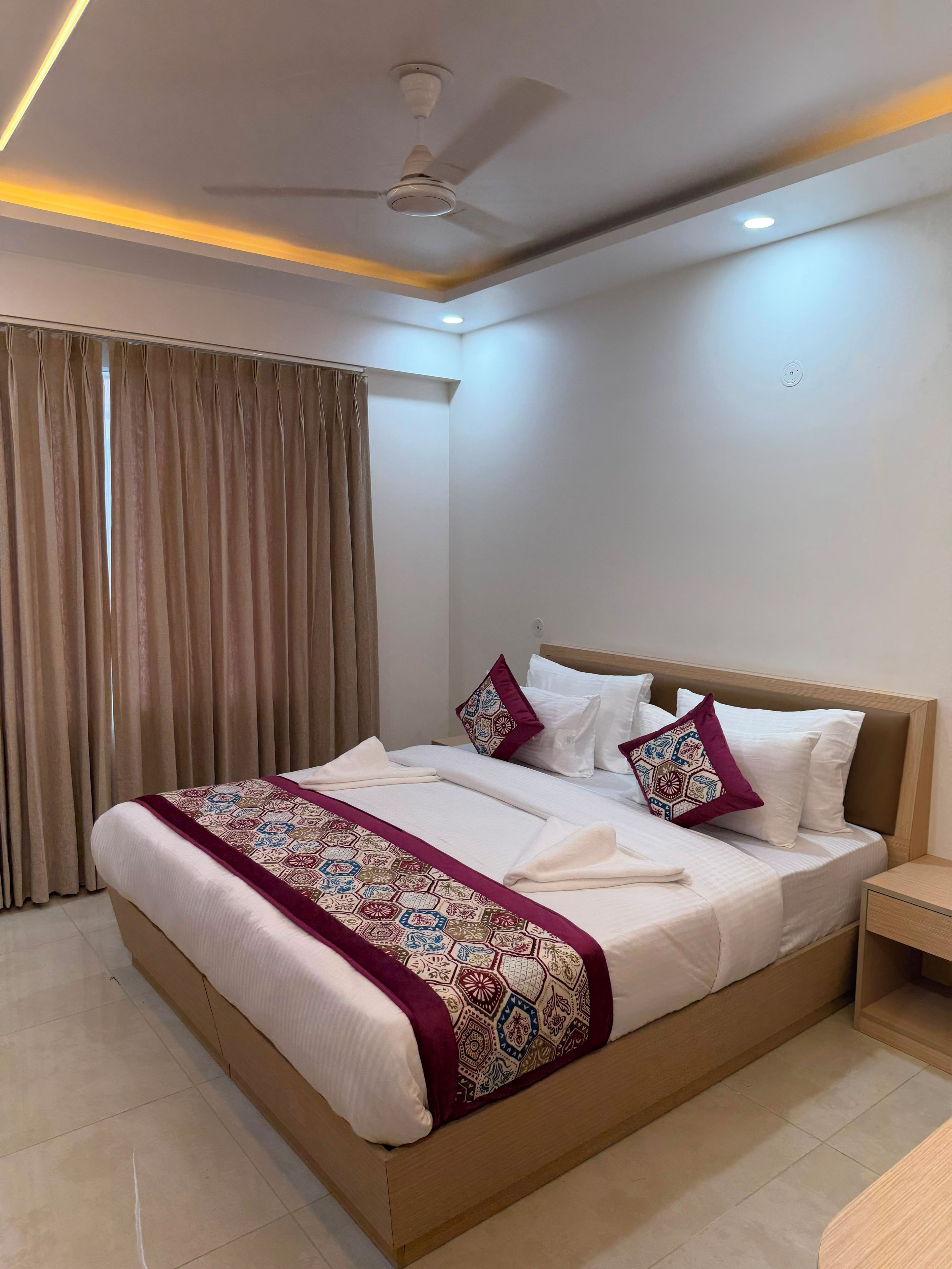 A hotel room with a bed, curtains, a ceiling fan, and a nightstand.
