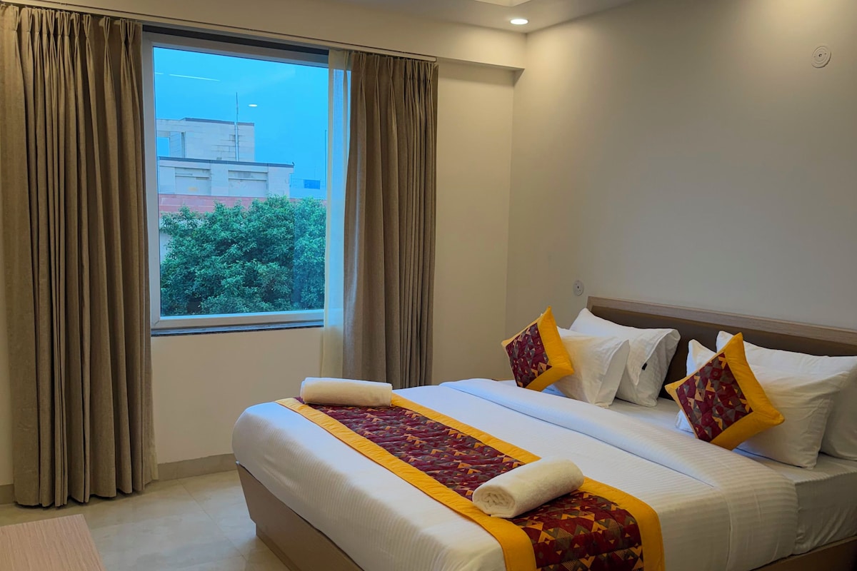 A hotel room with a large bed, a ceiling fan, a window with curtains, and a view of a building outside.