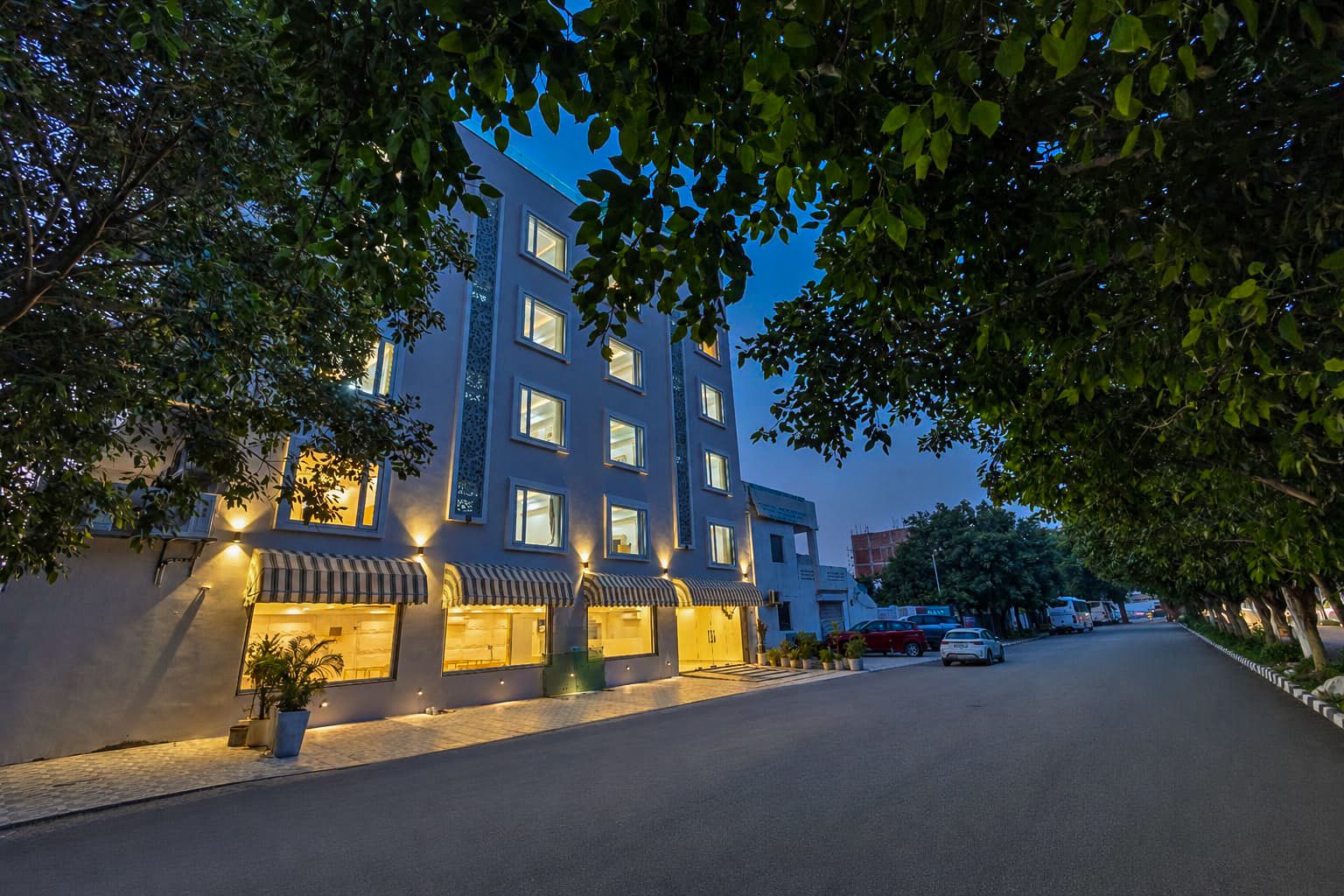 A multi-story hotel with a row of ground-floor shops, a paved street, and trees framing the scene.