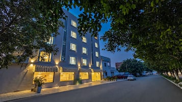A multi-story hotel with a row of ground-floor shops, a paved street, and trees framing the scene.