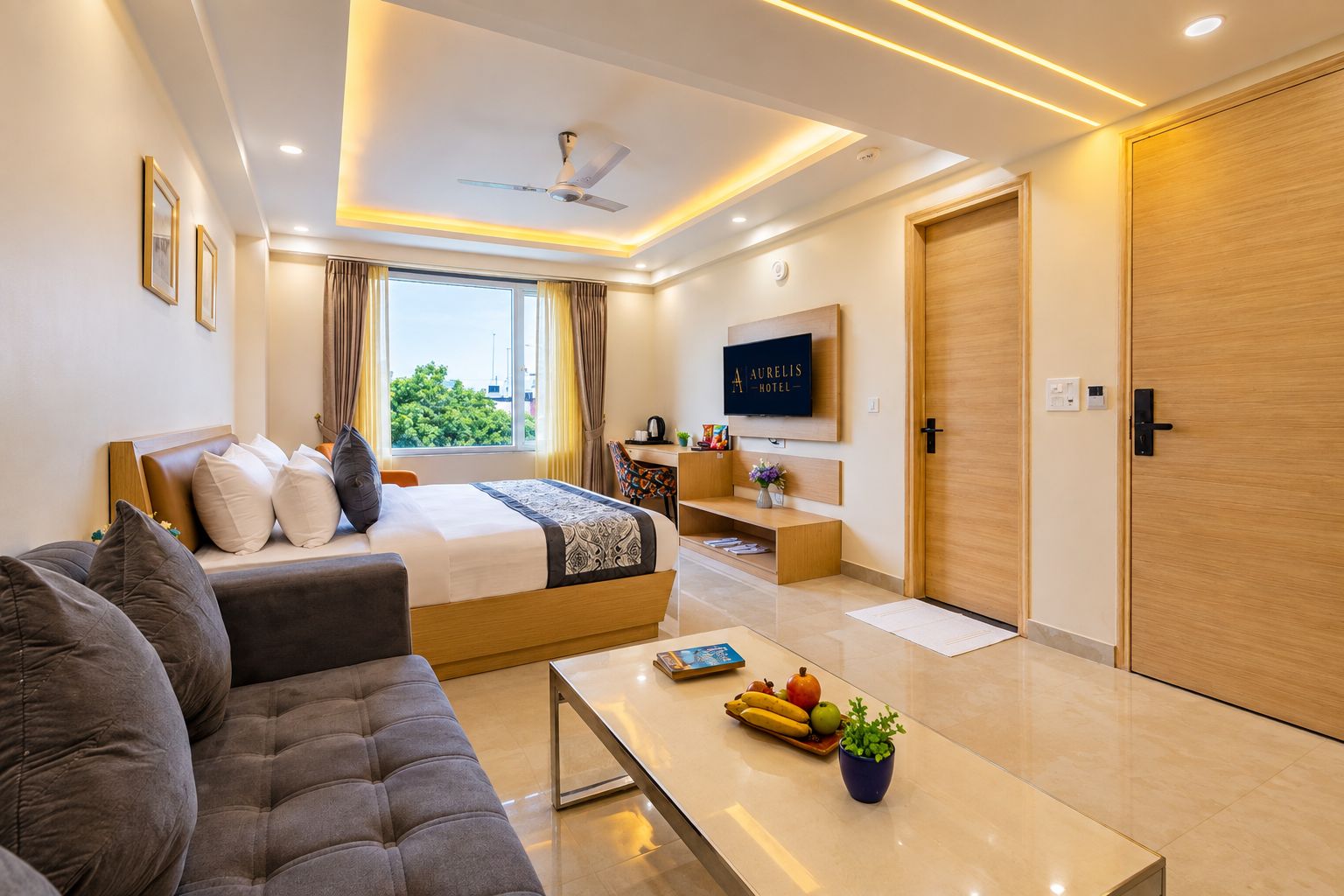A modern hotel room with a bed, sofa, TV, and a dining area with fruits and a plant.