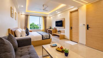 A modern hotel room with a bed, sofa, TV, and a dining area with fruits and a plant.