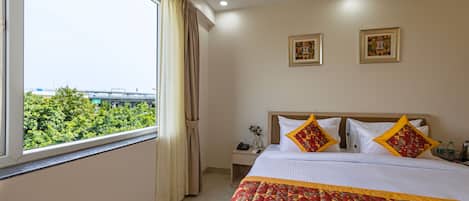 Quarto de hotel com uma cama grande, duas obras de arte emolduradas na parede, uma mesa de cabeceira com telefone e um vaso, e uma janela com cortinas.