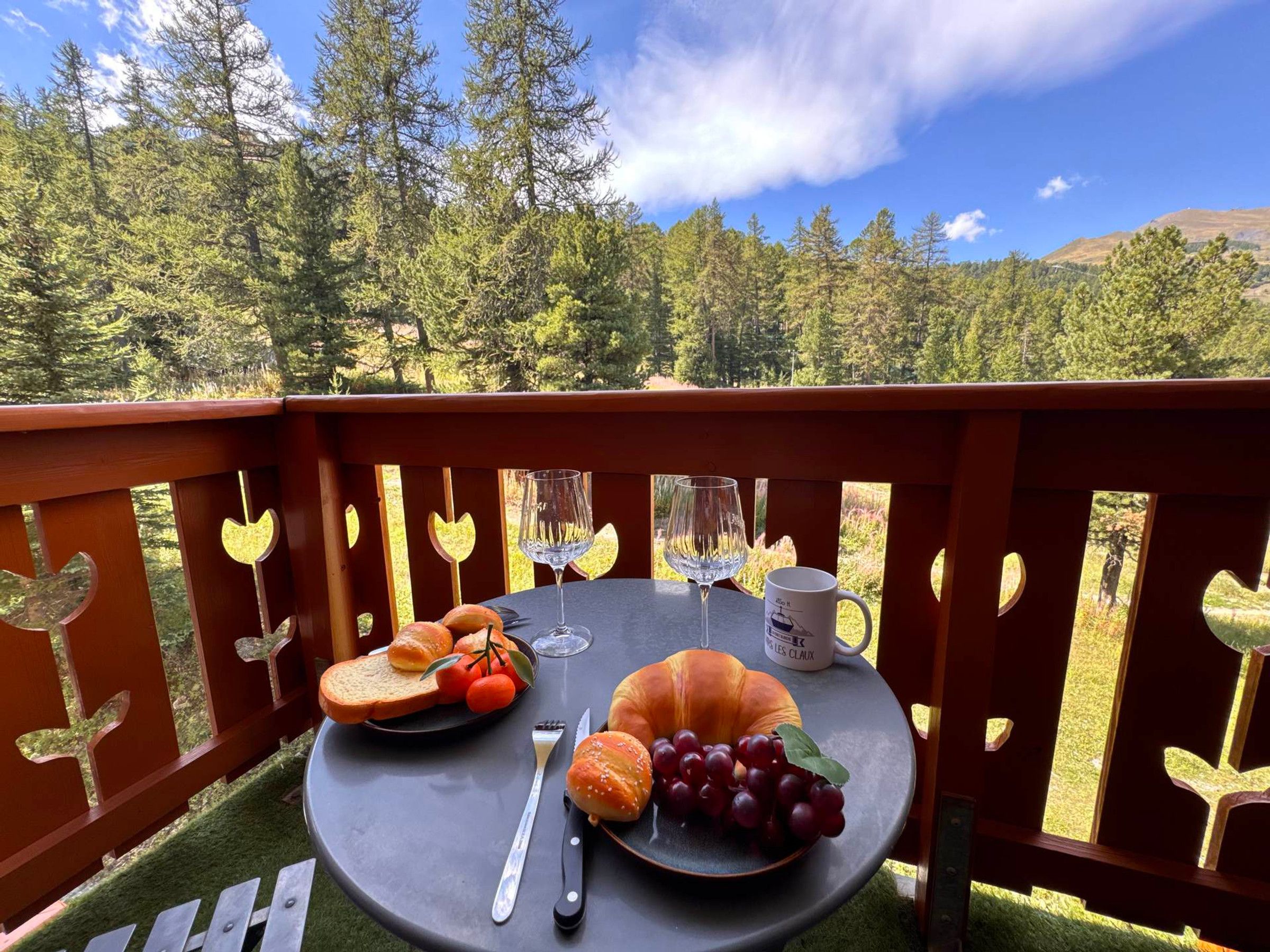 A table set with bread, grapes, a croissant, a wine glass, and a mug on a balcony with a scenic forest view.