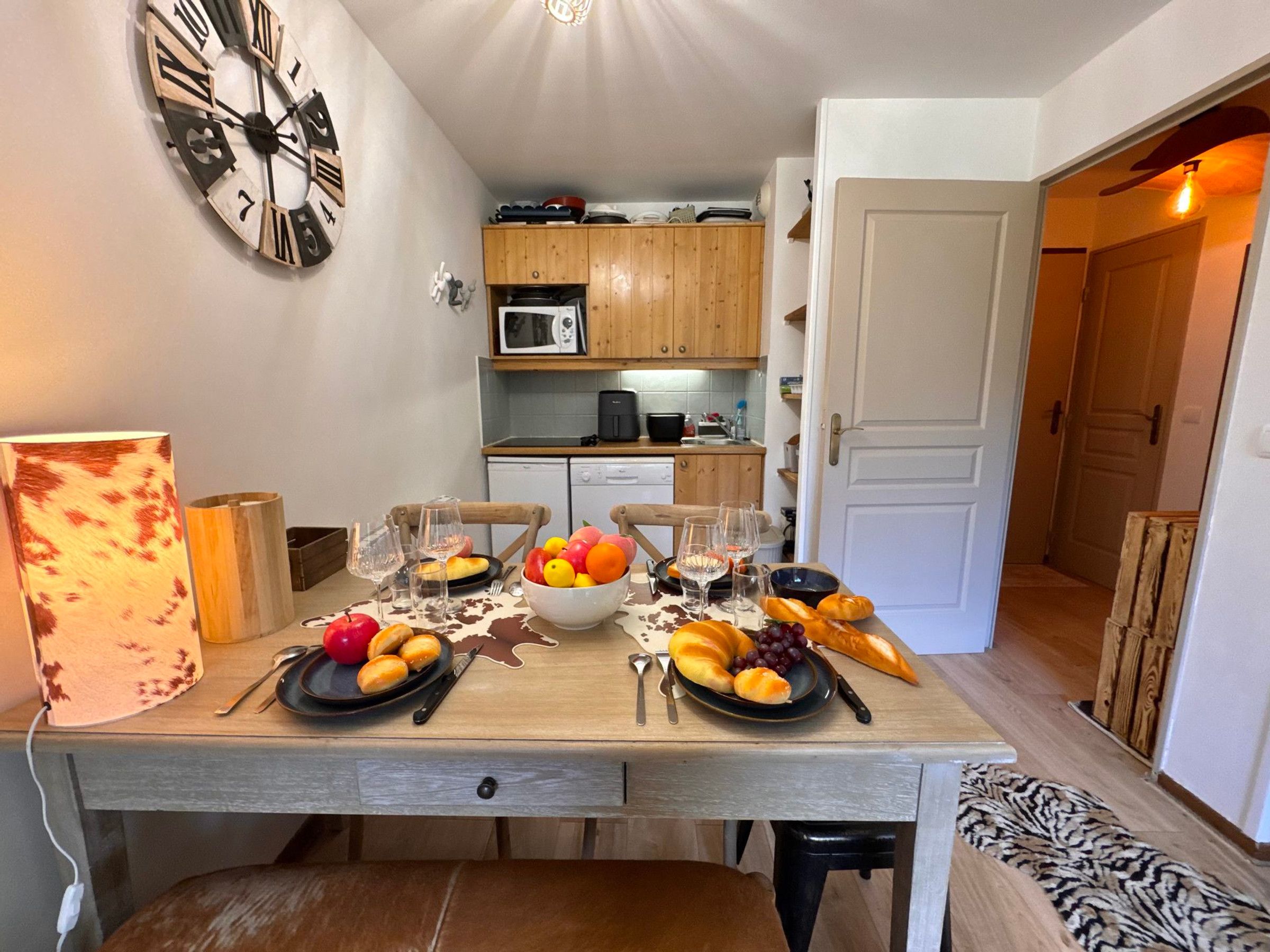 A dining table set with plates of fruit, wine glasses, and bread, with a clock on the wall and a kitchen in the background.