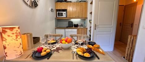 Una mesa de comedor dispuesta con platos de fruta, copas de vino y pan, con un reloj en la pared y una cocina al fondo.