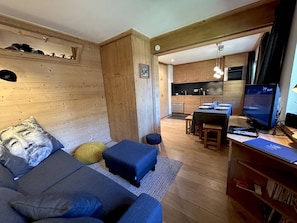 Ein gemütliches Wohnzimmer mit einer Couch, Poufs und einem Fernseher. Im Hintergrund ist eine Küche zu sehen.