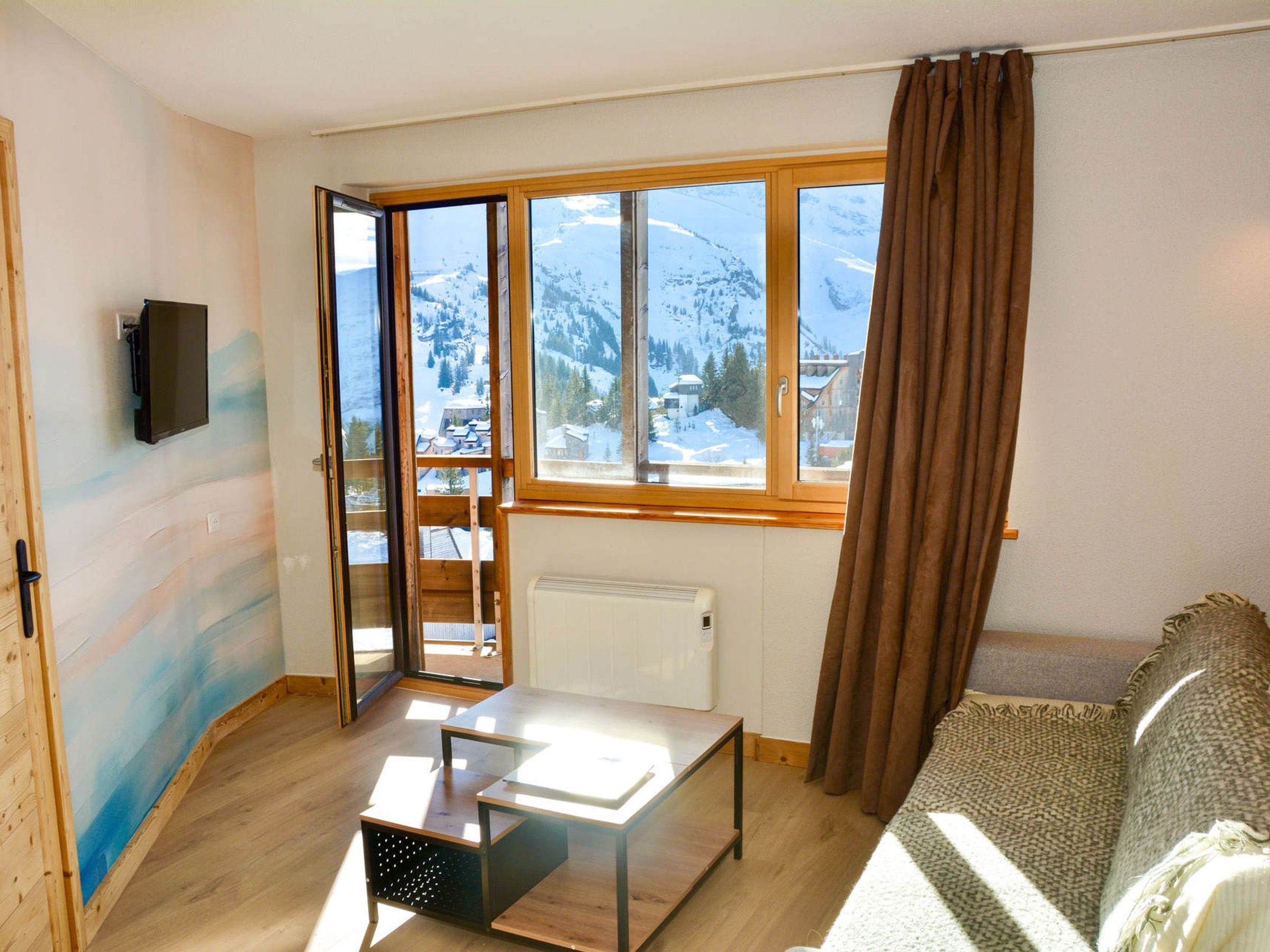 Una habitación con suelo de madera, una mesa, un sofá, una ventana que ofrece vistas a montañas nevadas y un radiador.
